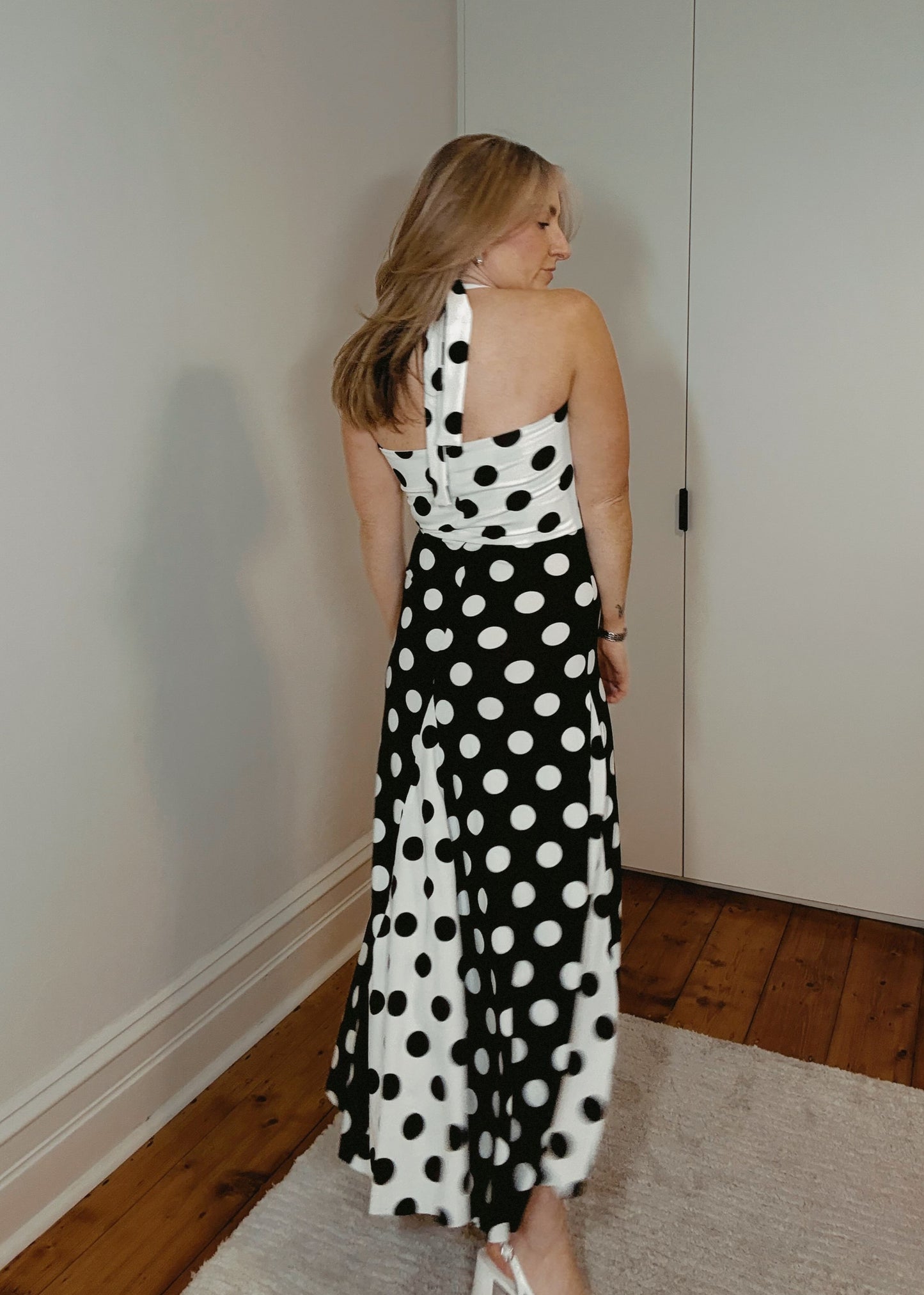 Woman in a black and white polka dot dress standing in a room with wooden flooring and a white wardrobe.