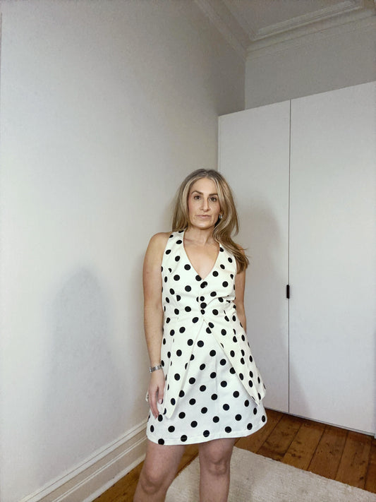Woman wearing a polka dot dress standing in a room with white walls and wooden floor.
