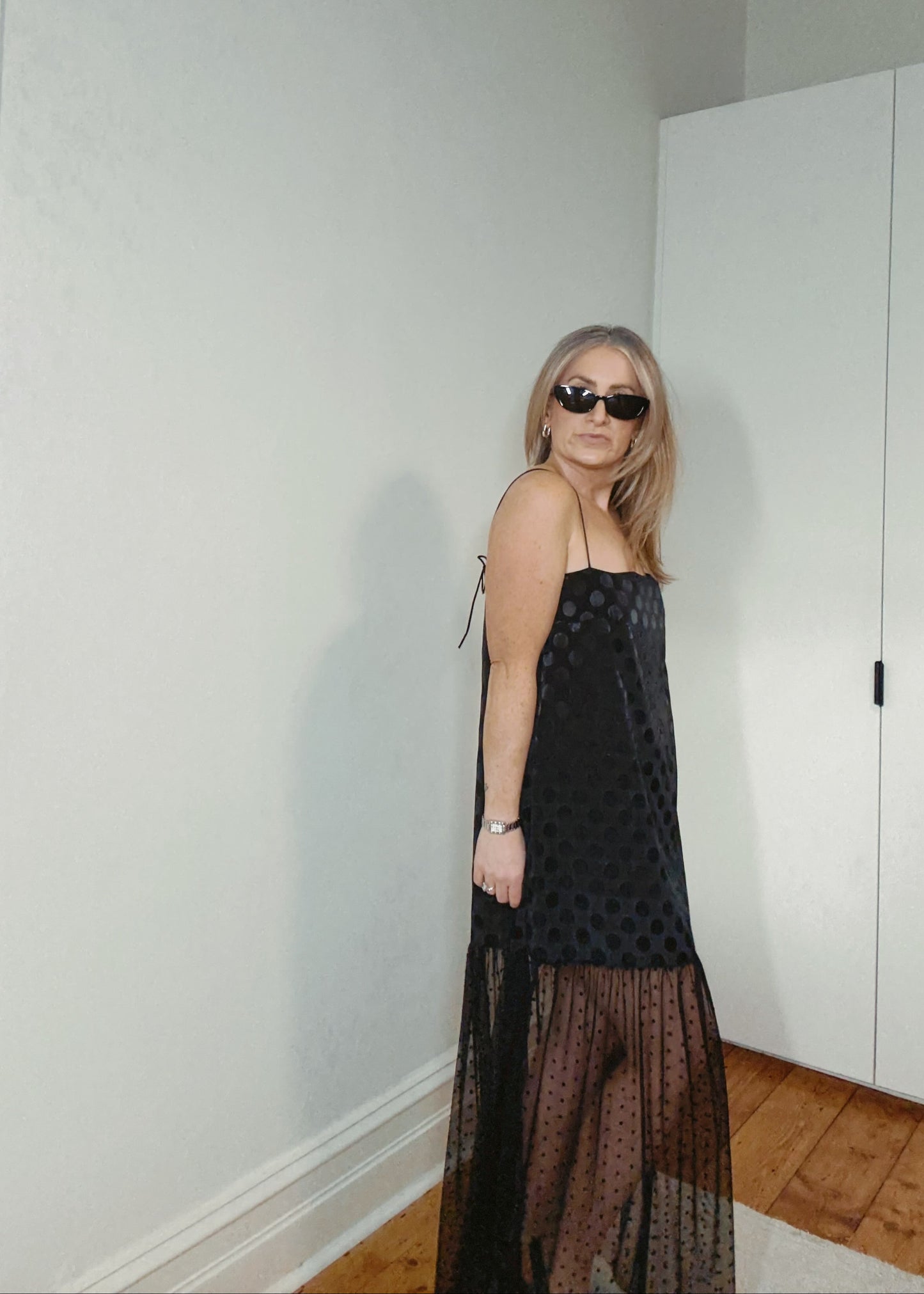 Person wearing a black dress with sheer overlay in a room with white walls and wooden floor.