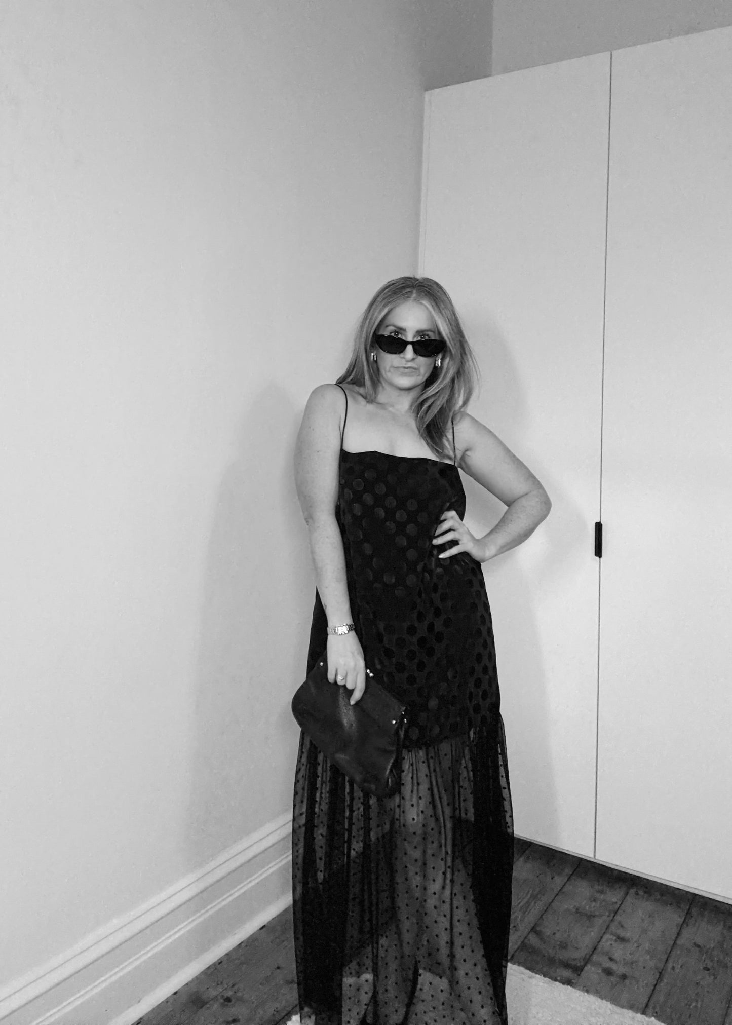 Person wearing a black dress and sunglasses in a room with white walls and wooden flooring.