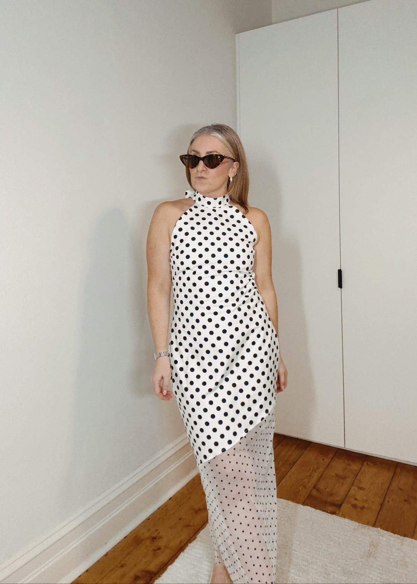 Woman wearing a black and white polka dot dress in a room with white walls and wooden flooring.