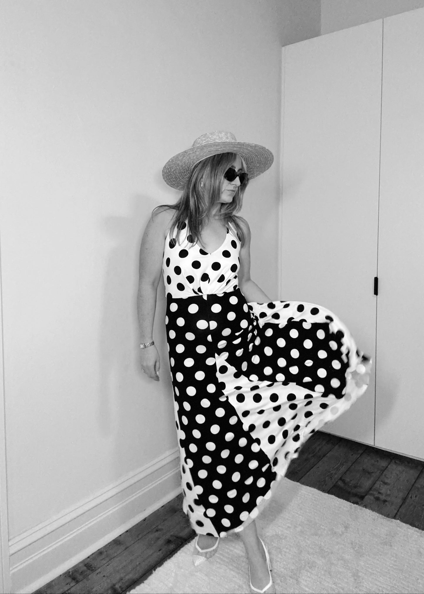 Woman in a polka dot dress and hat standing in a room with white walls and wooden floor.