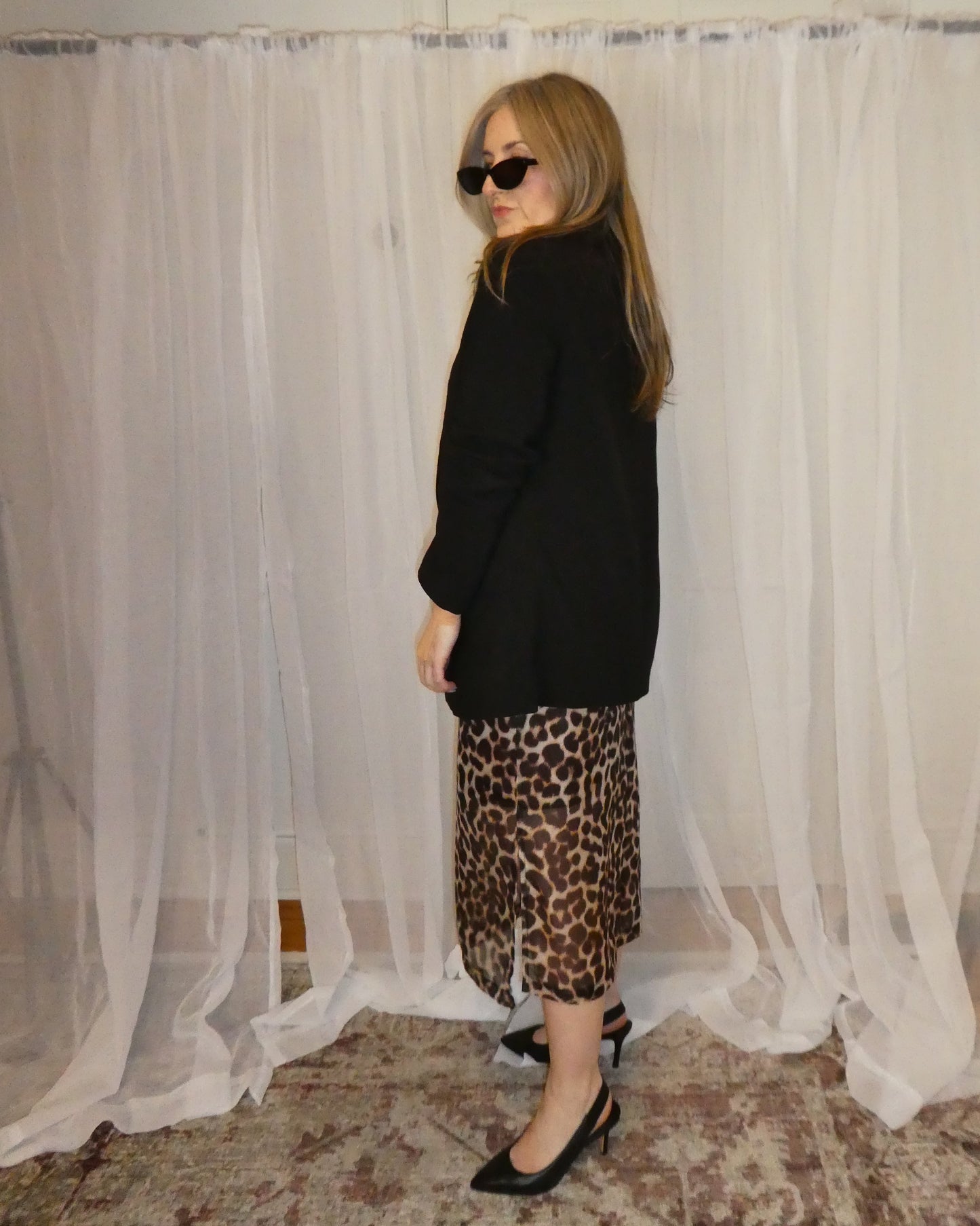 Person wearing a black top and leopard print skirt standing in front of a white curtain.