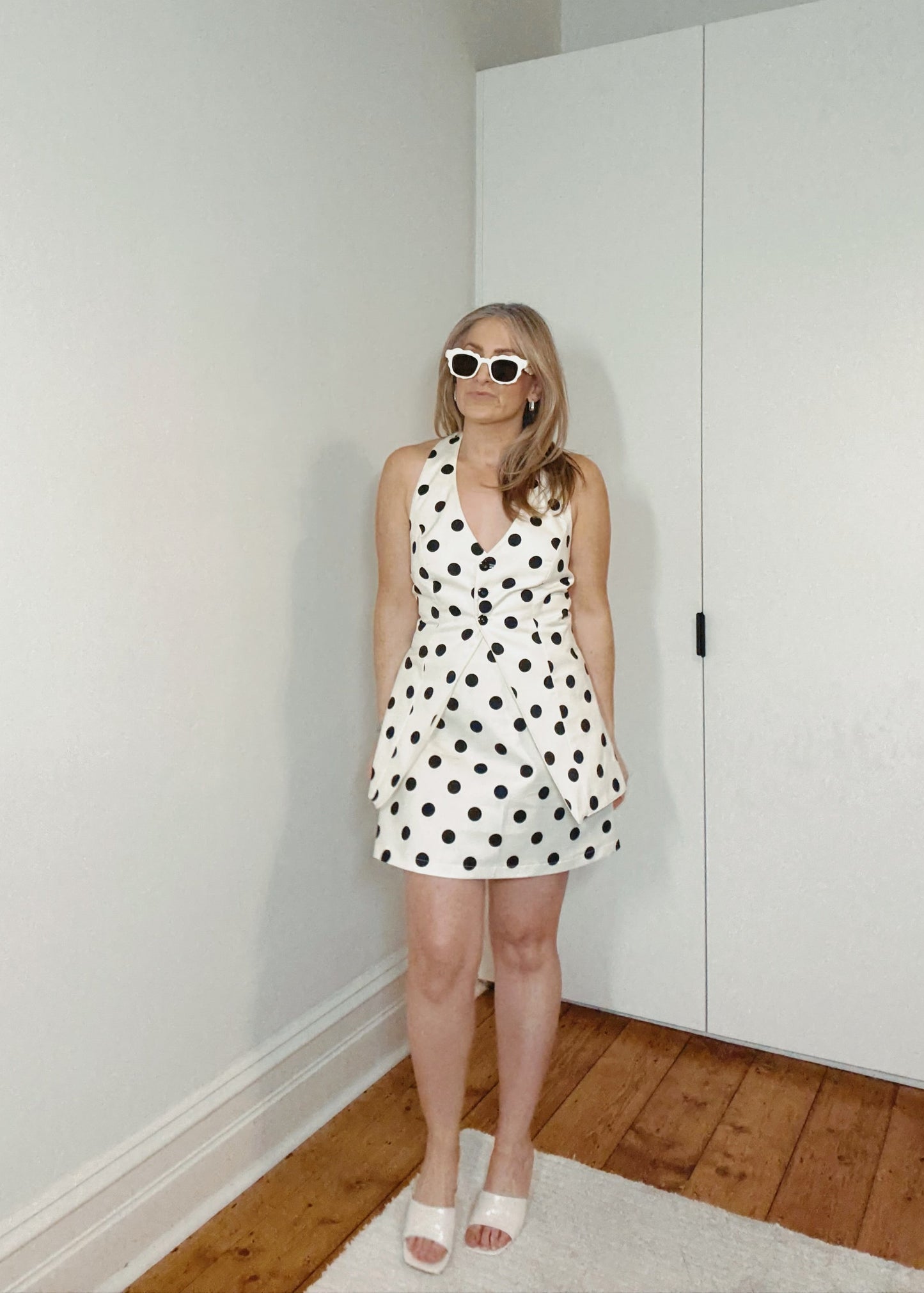 Woman in a polka dot dress standing in a room with white walls and wooden flooring.