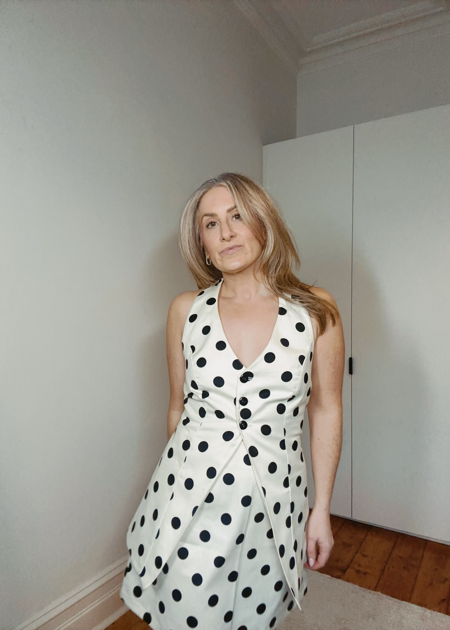 Woman wearing a white dress with black polka dots in a room with a white wall and wooden floor.