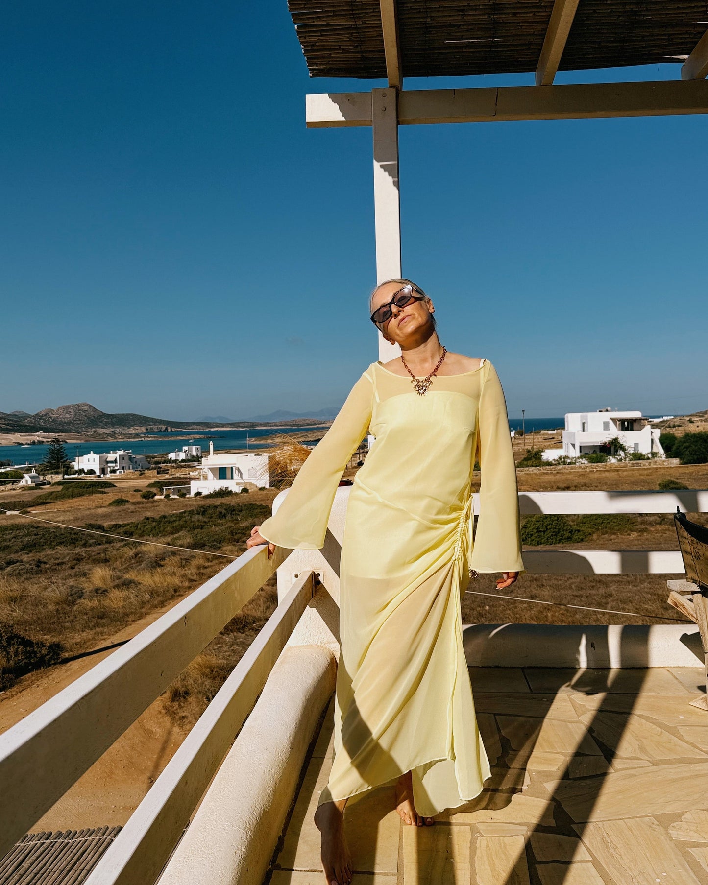 Woman in a light yellow dress standing on a balcony with a scenic view of the sea and mountains.