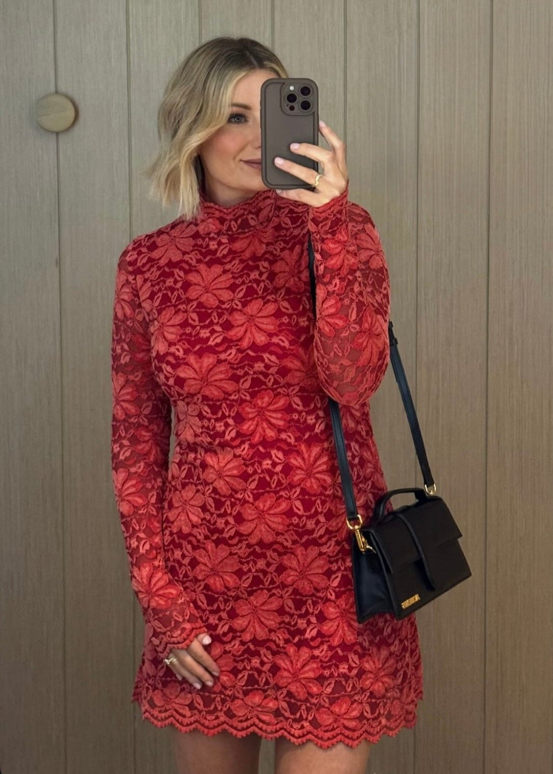 Person wearing a red dress with a pattern, holding a phone and a black handbag against a wooden wall.