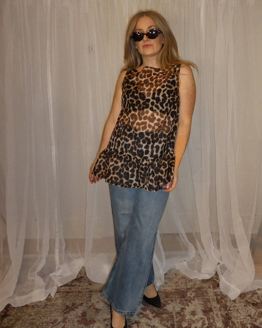 Woman wearing a leopard print top and blue jeans standing in front of a white curtain.