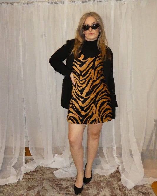 Person wearing a zebra print dress and black coat in front of sheer curtains