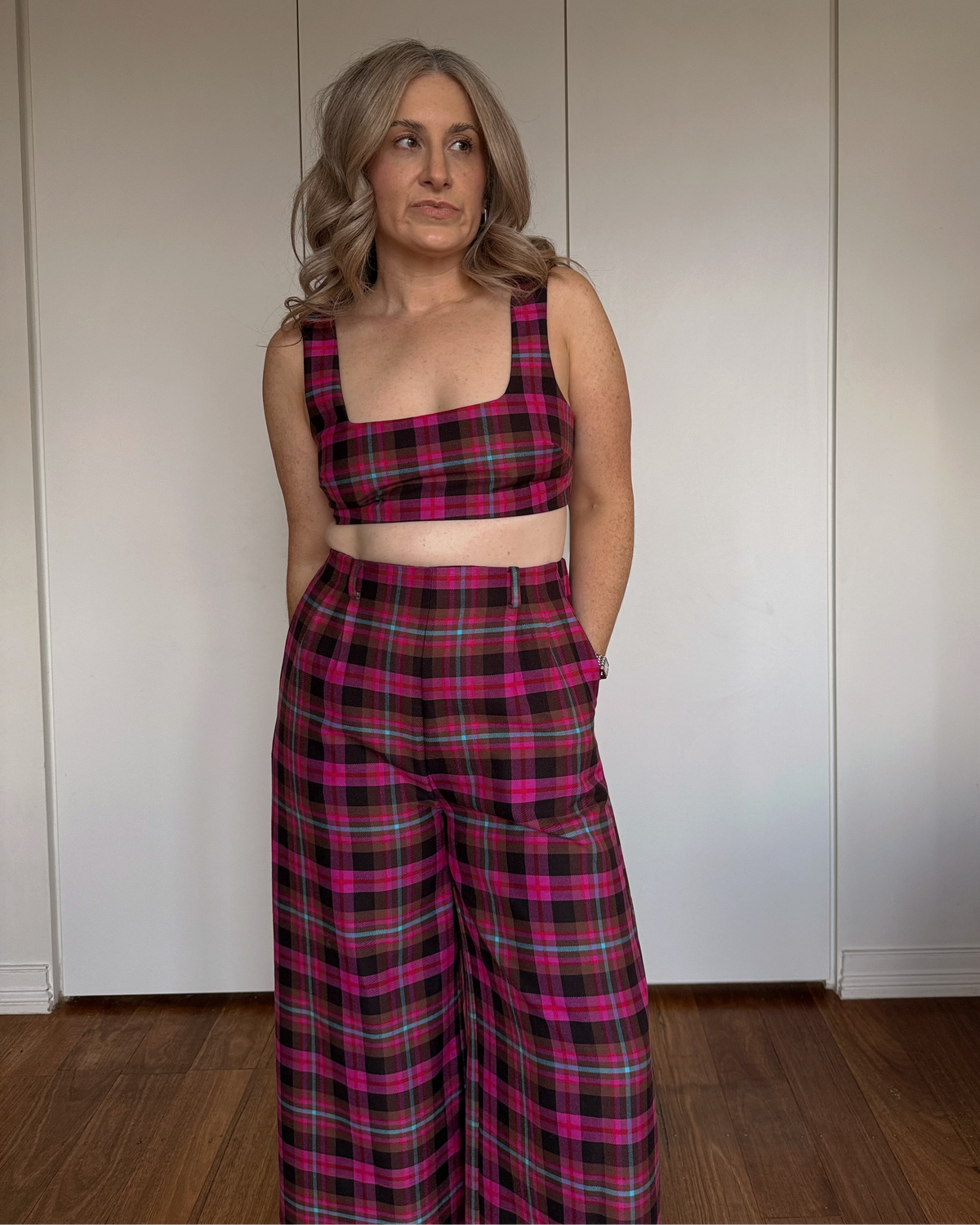 A woman stands indoors wearing a matching two-piece plaid outfit with a cropped tank top and wide-leg trousers in a pink tartan pattern