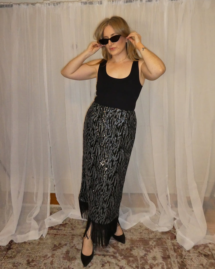Woman wearing a black top and patterned skirt standing in front of sheer curtains.