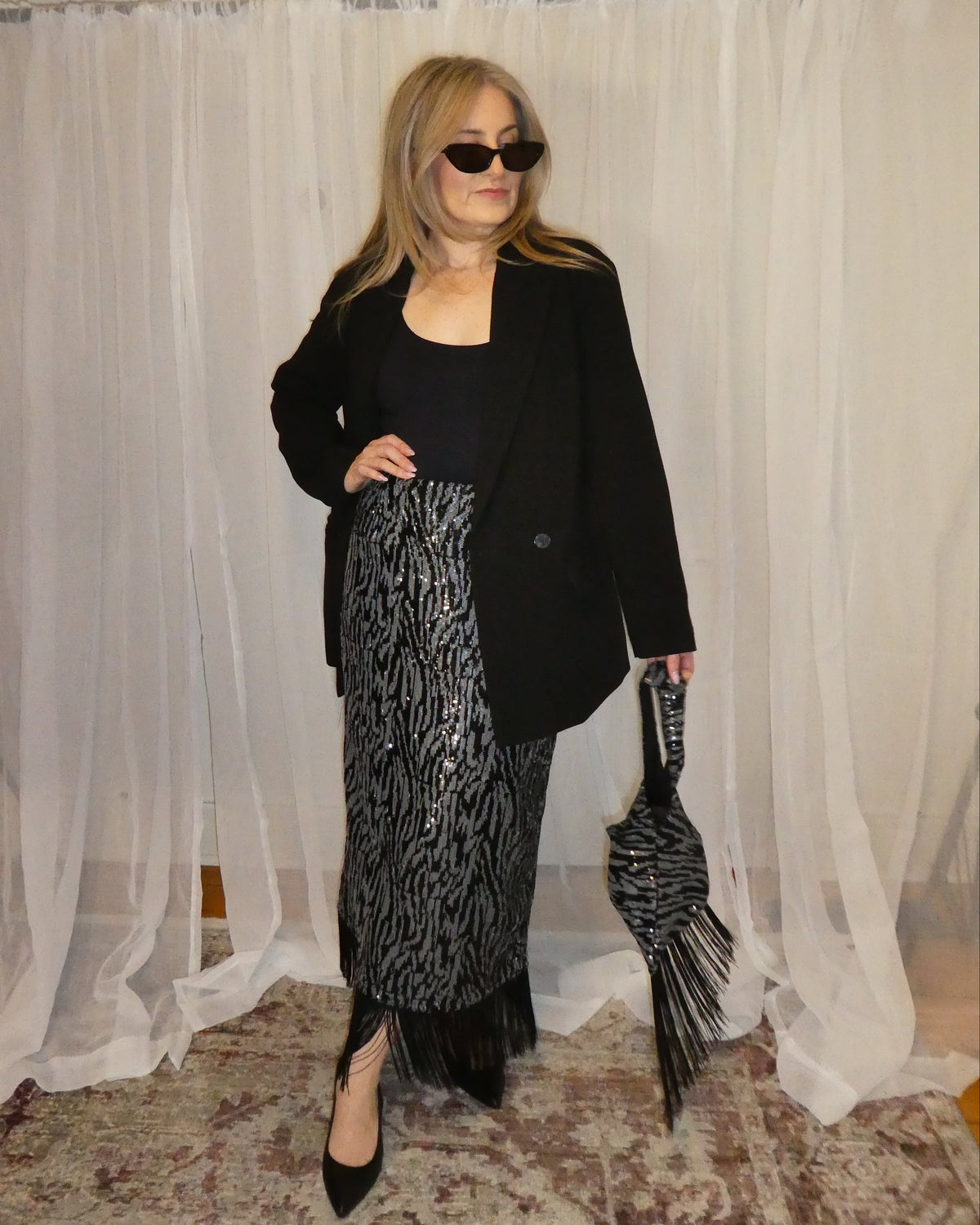 Person wearing a black outfit with a patterned skirt in front of sheer curtains.