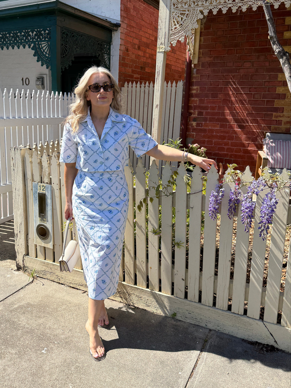 A woman wearing a light blue and white patterned short-sleeve shirt dress stands on a sidewalk in front of a white picket fence and brick house.