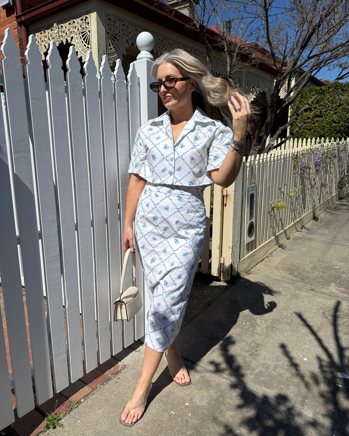 A woman wearing a matching light blue and white patterned short-sleeve shirt and midi skirt set, walking barefoot on a sidewalk next to a white picket fence.