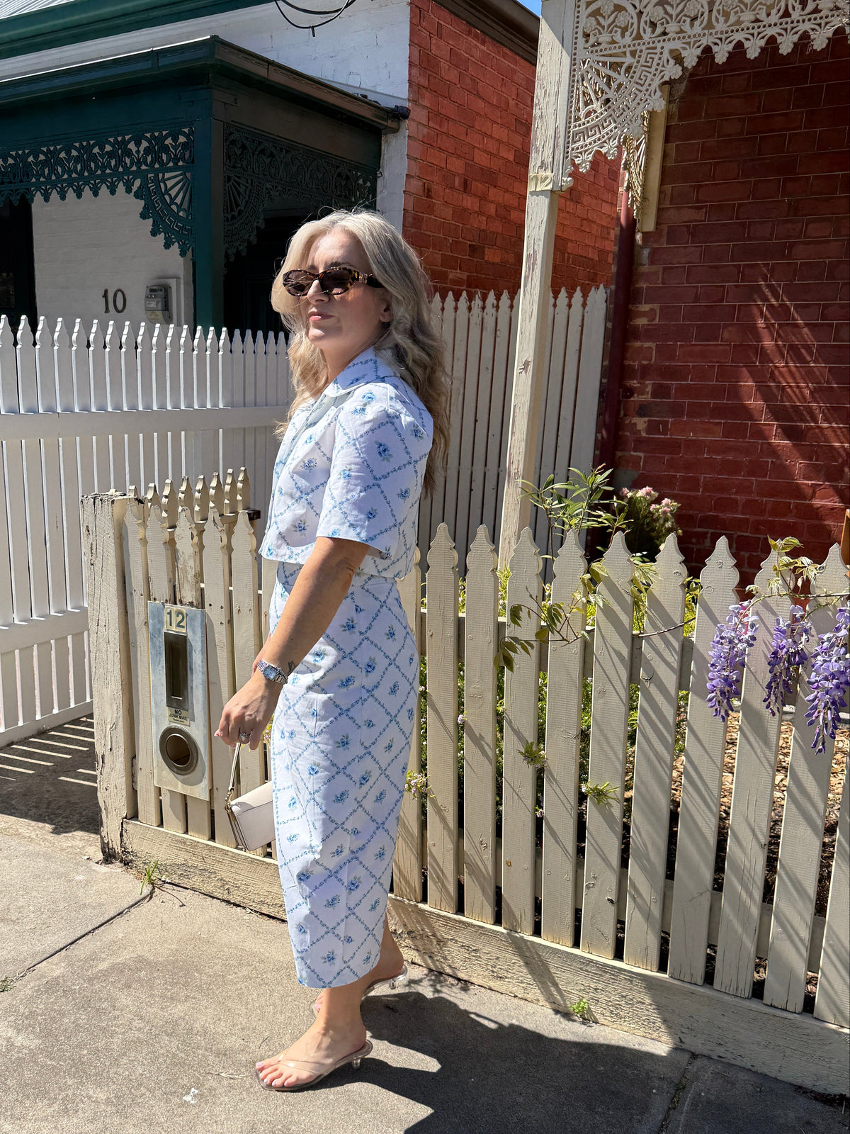 A woman wearing a light blue and white patterned short-sleeve crop top and matching midi skirt stands barefoot on a sidewalk in front of a white picket fence and brick house.