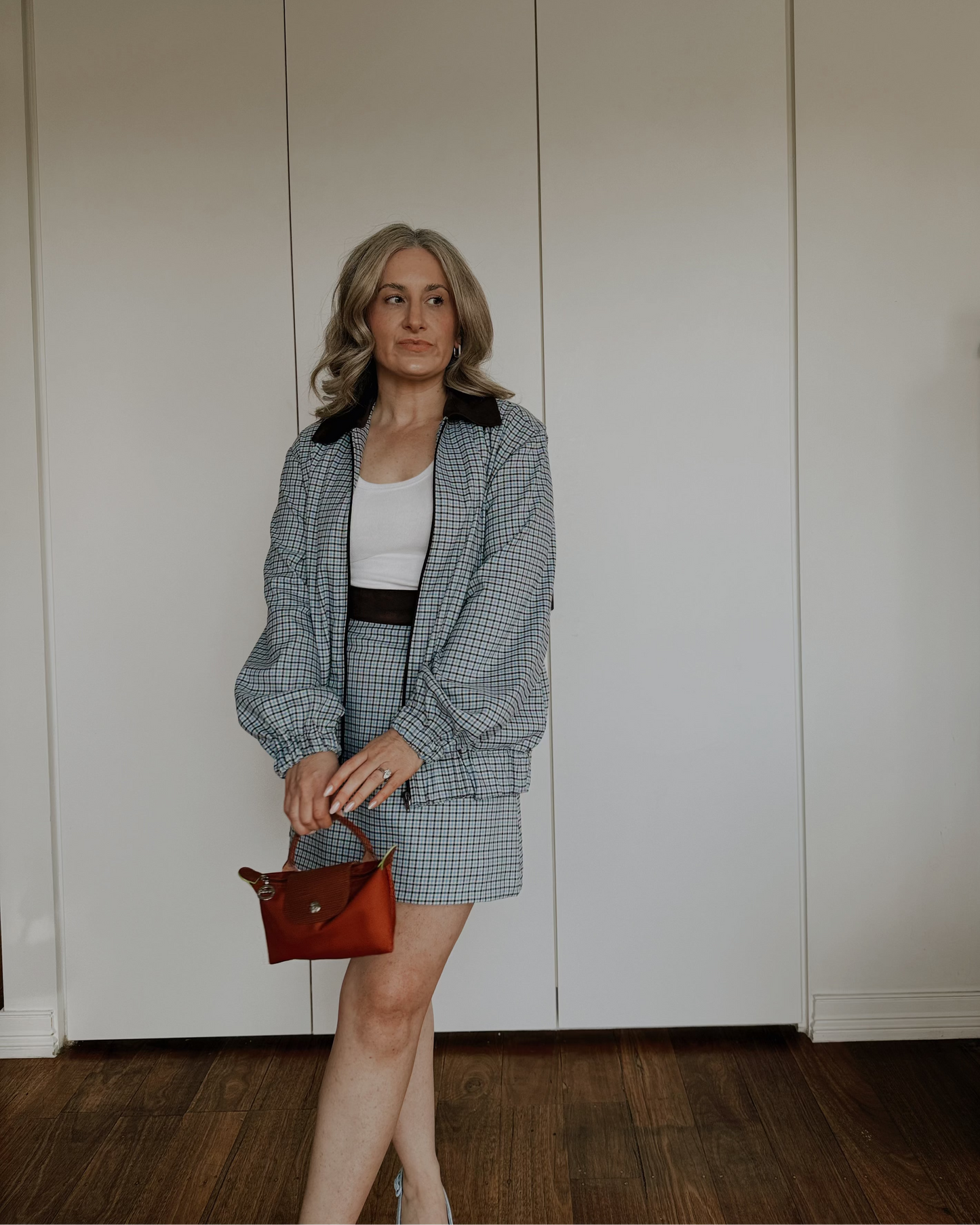 A woman stands in a room with white closet doors and dark wood flooring, wearing a blue and white gingham blazer and matching mini skirt over a white top, holding a small brown handbag.