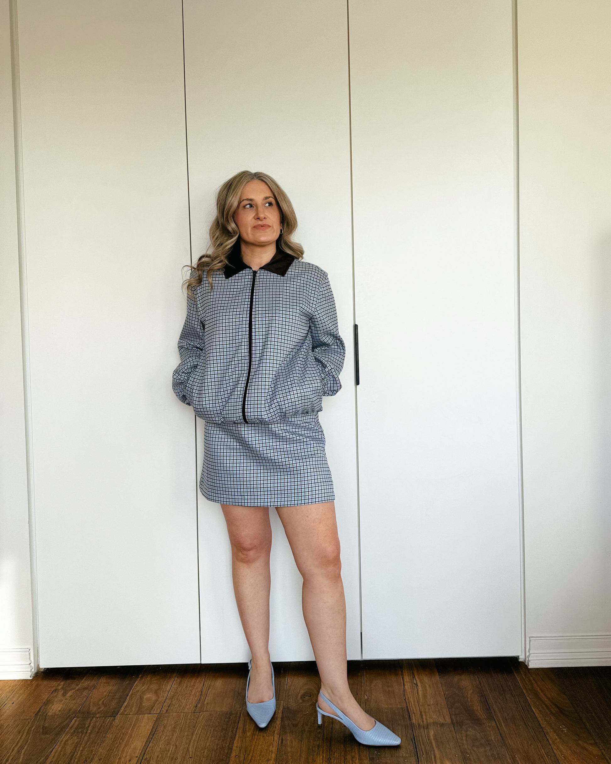 A woman stands in front of a white wardrobe door wearing a matching light blue and white gingham mini skirt and jacket set with a brown collar, paired with light blue pointed-toe heels.
