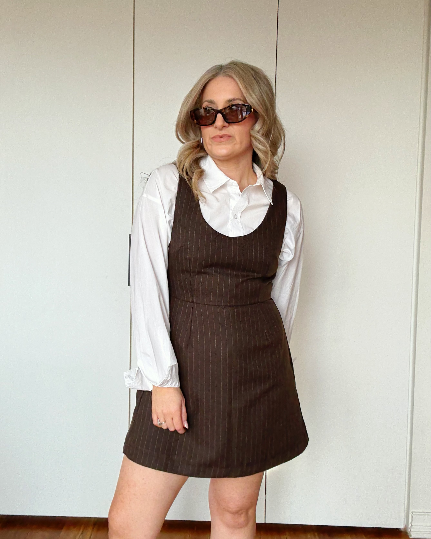 A woman wearing a brown pinstriped dress over a white button-down shirt, styled with dark sunglasses.