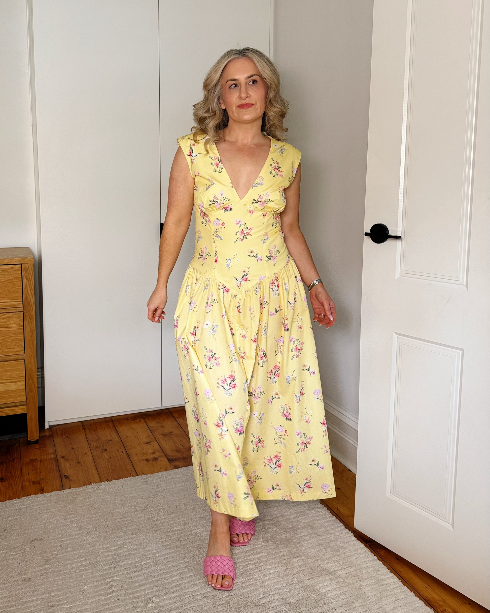 A woman wearing a yellow sleeveless maxi dress with a pink and green floral print stands in a bedroom doorway, wearing pink slip-on sandals.