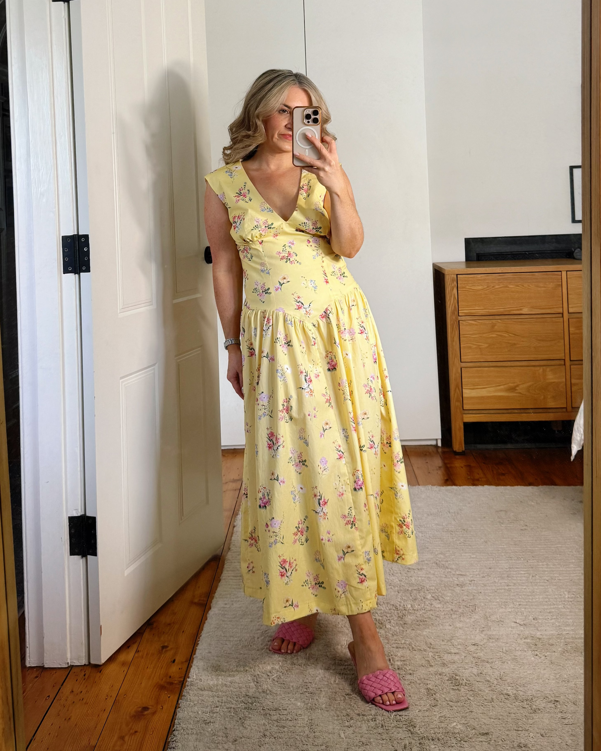 A woman wearing a yellow floral midi dress and pink shoes stands in a bedroom taking a mirror selfie. The image is framed by a large wooden doorway.