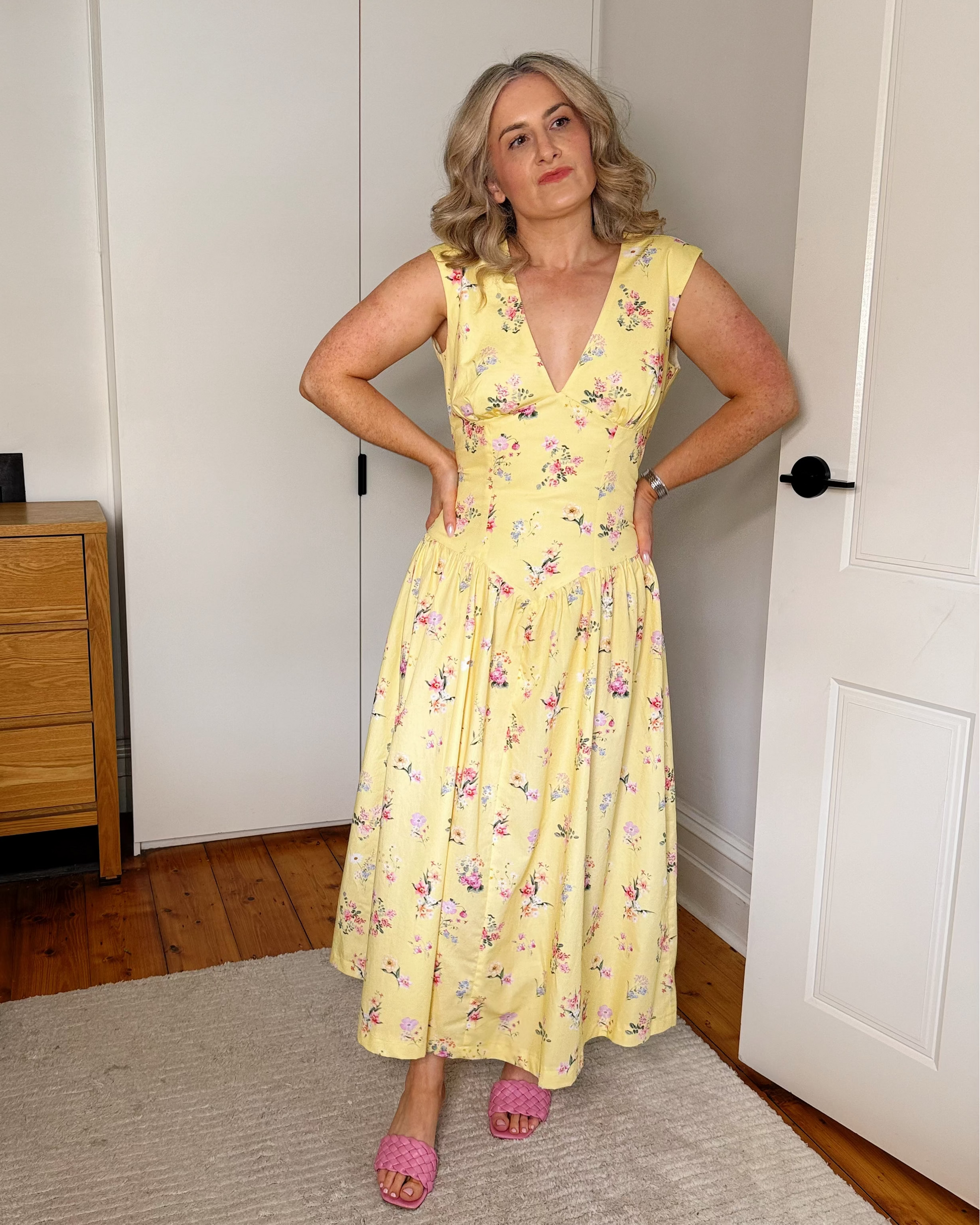 A woman stands in a bedroom wearing a sleeveless yellow maxi dress with a pink and green floral print, paired with pink sandals.