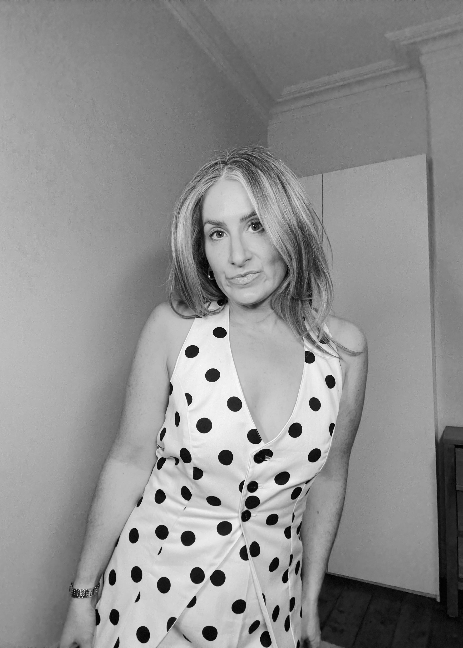 Woman wearing a polka dot dress in a room with a plain wall and wooden floor.