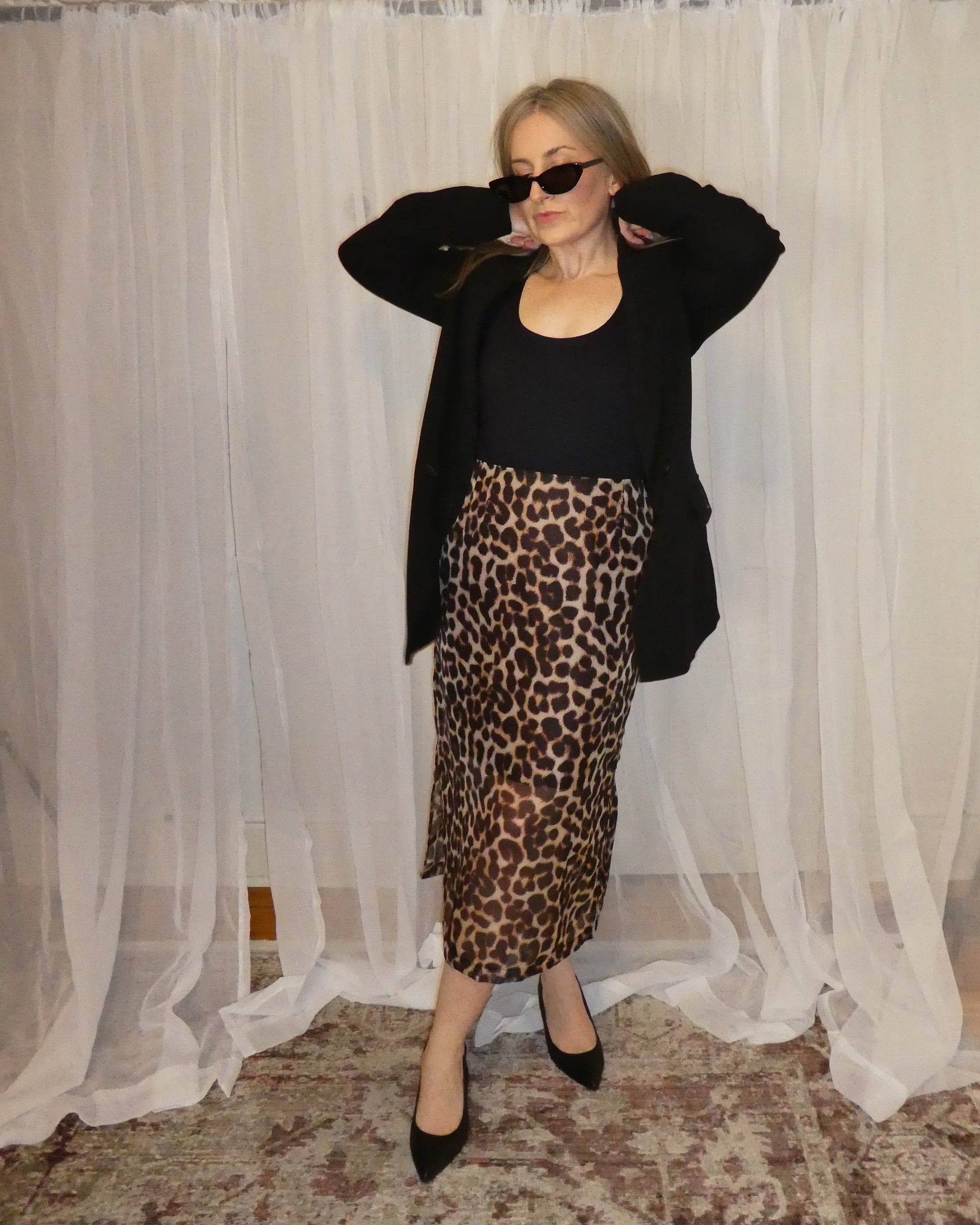 Woman wearing a black top and leopard print skirt in front of sheer curtains.