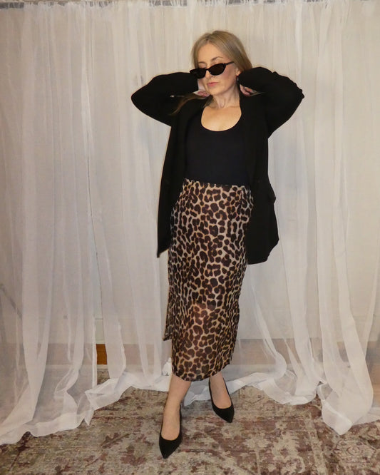 Woman wearing a black top and leopard print skirt in front of sheer curtains.