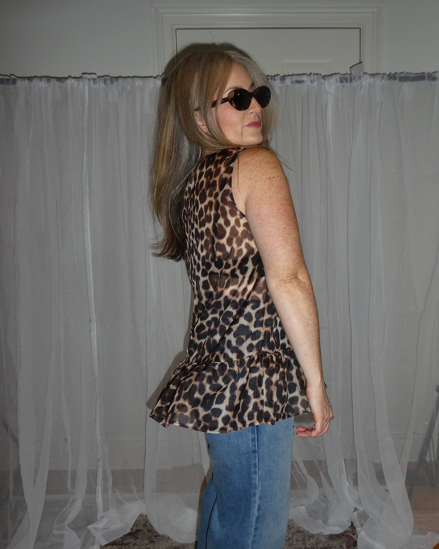 Person wearing a leopard print top and blue jeans standing in front of sheer curtains.