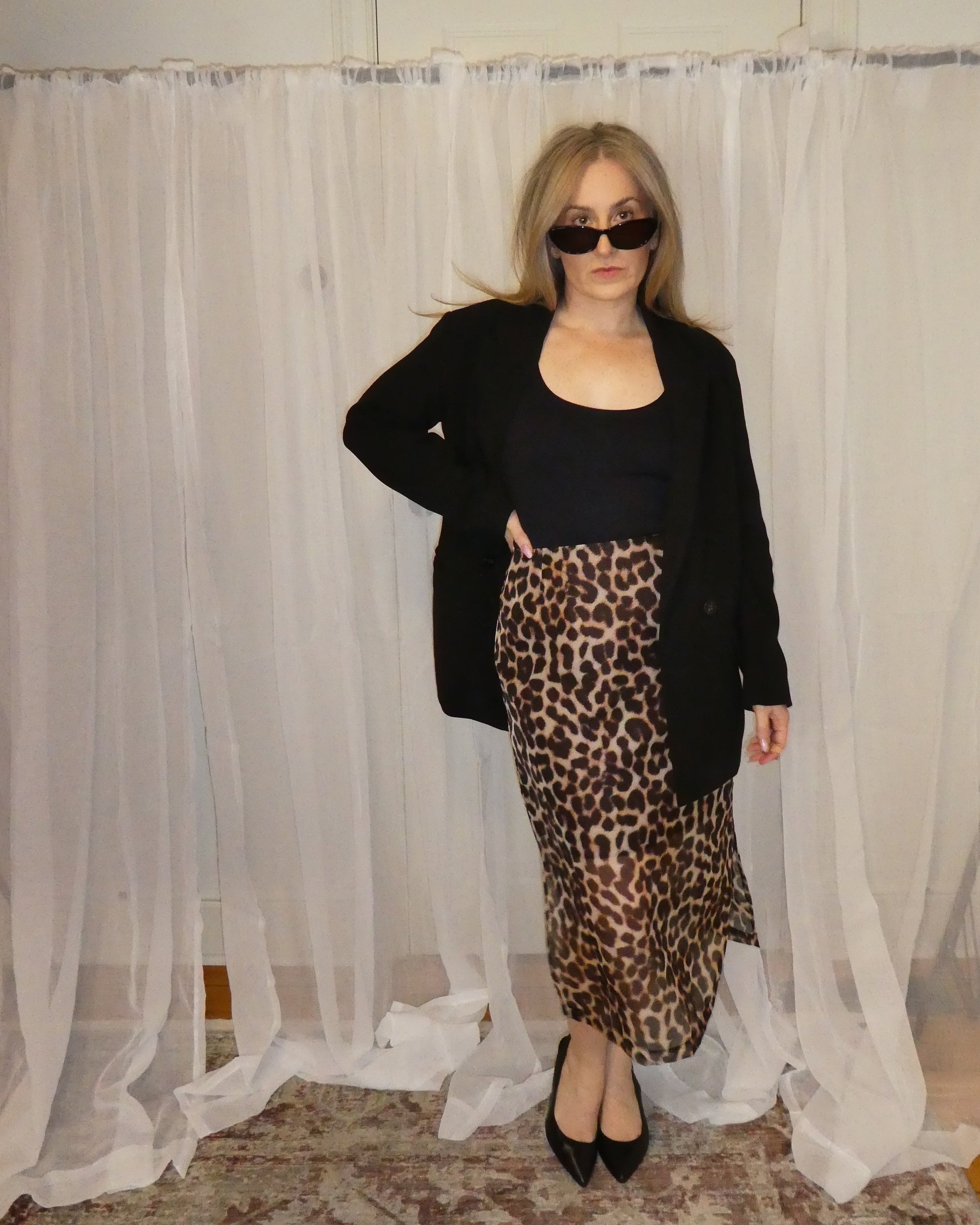 Person wearing a black cardigan, black top, and leopard print skirt standing in front of sheer curtains.