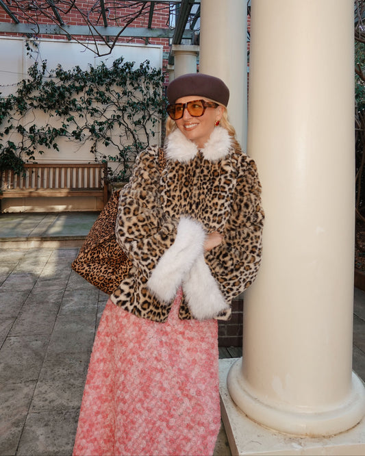 A woman wearing a leopard print faux fur coat with white fur trim, a brown beret, orange-tinted sunglasses, and a pink floral skirt, standing in an outdoor patio setting with large cream columns.