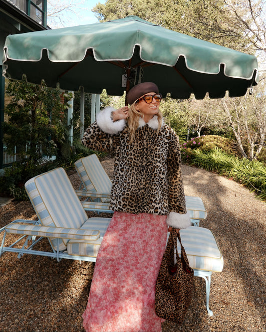 A woman stands outdoors under a large green patio umbrella wearing a leopard-print jacket with white fur trim, a pink patterned skirt, and a brown hat. She is posed in front of lounge chairs and trees on a sunny day.