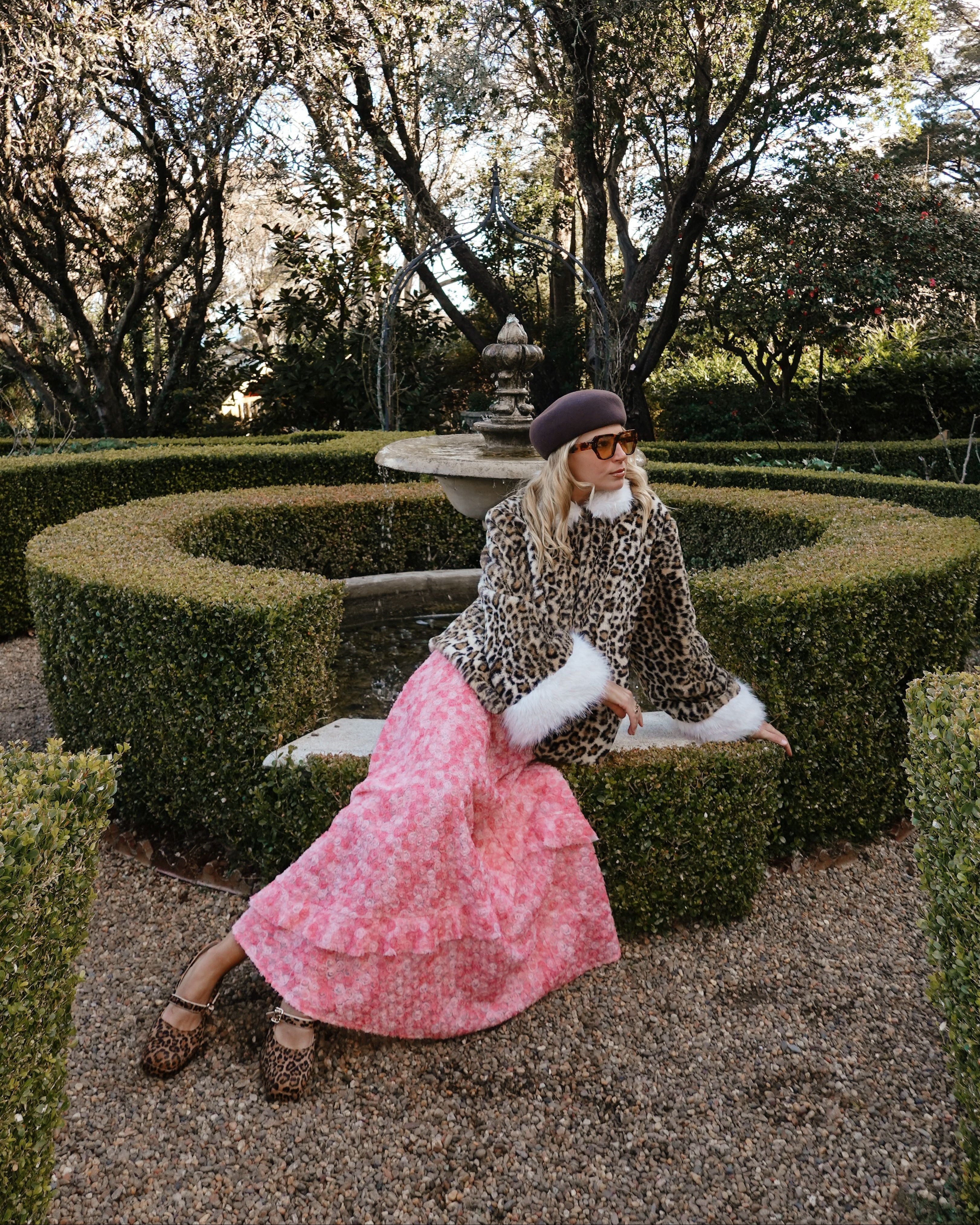 A woman posing in a formal garden setting, wearing a leopard-print jacket with white fur trim, a pink tiered floral skirt, and a dark purple beret.