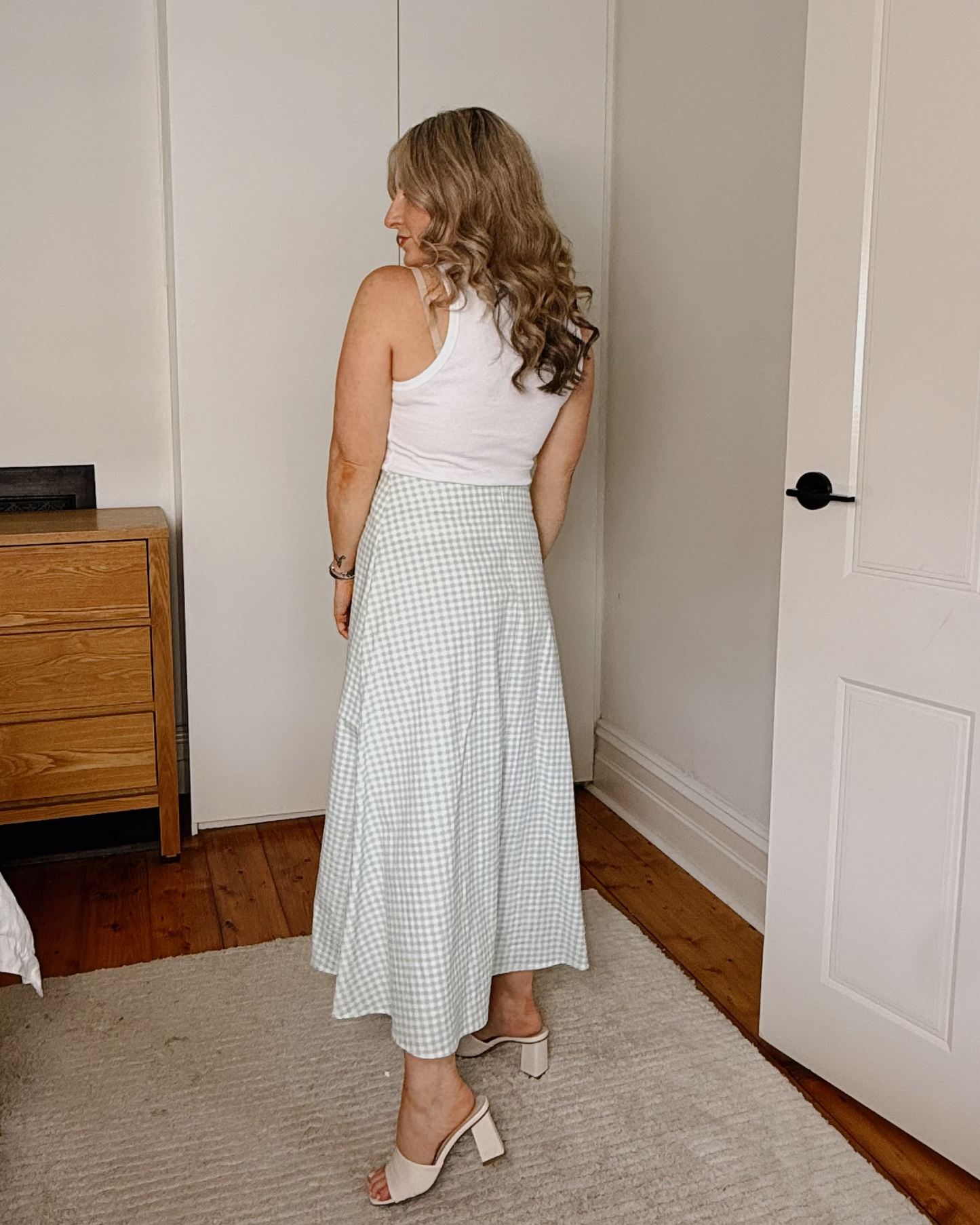 A woman stands in a bedroom wearing a sleeveless white top and a light green and white gingham midi skirt, paired with white heeled sandals.