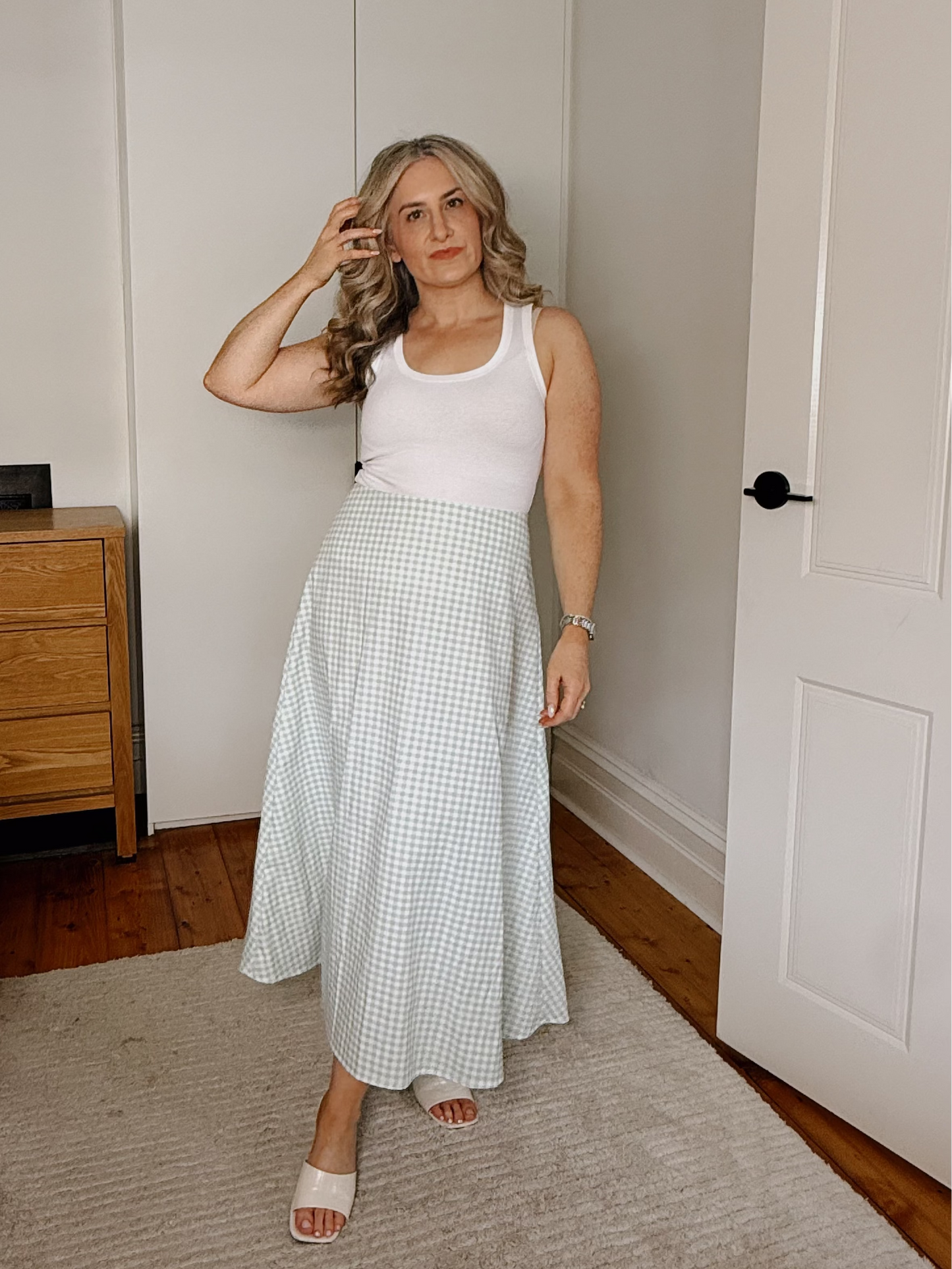 A woman stands in a bedroom wearing a sleeveless white tank top and a light green and white gingham maxi skirt, paired with white slide sandals.