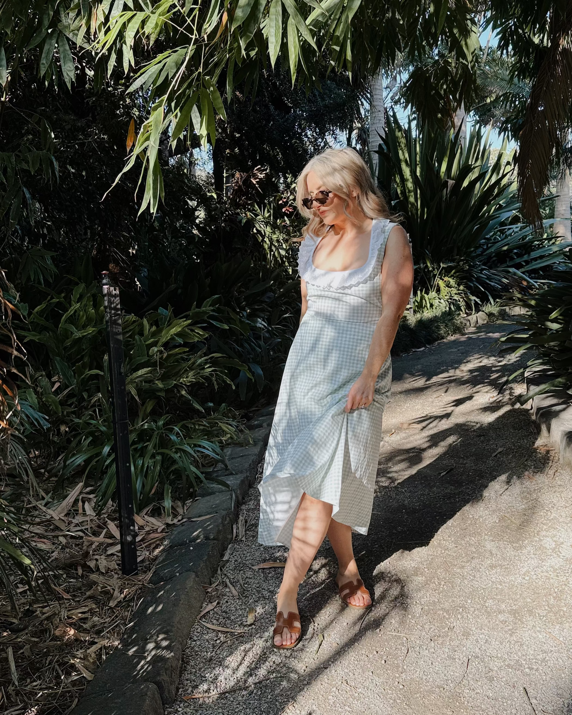 A woman wearing a light-colored sleeveless dress and sunglasses walks along a sun-dappled garden path surrounded by lush green foliage.