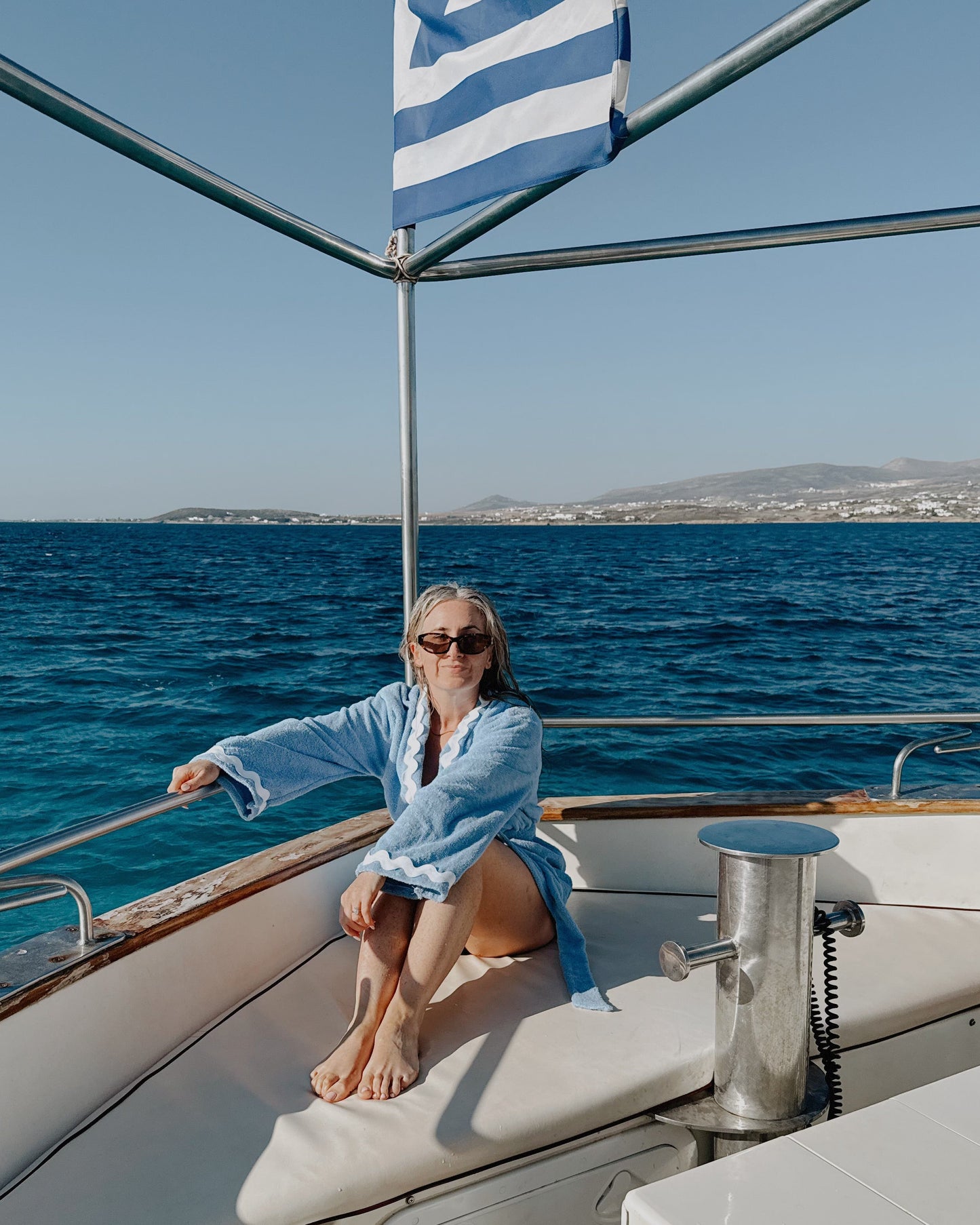 Woman sitting on a boat with a Greek flag, blue sea, and clear sky.