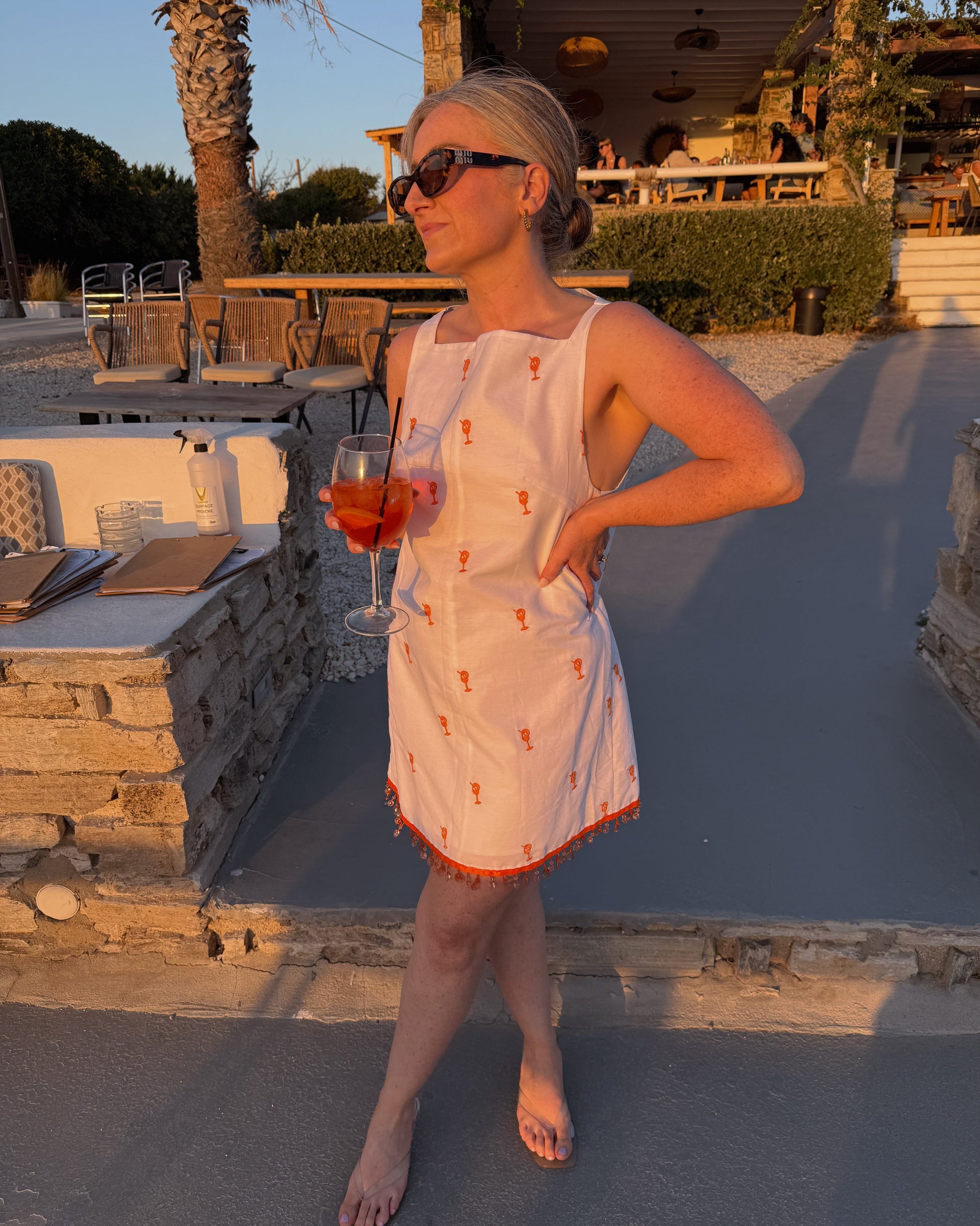 Woman in a white dress with orange patterns holding a glass of red wine outdoors.