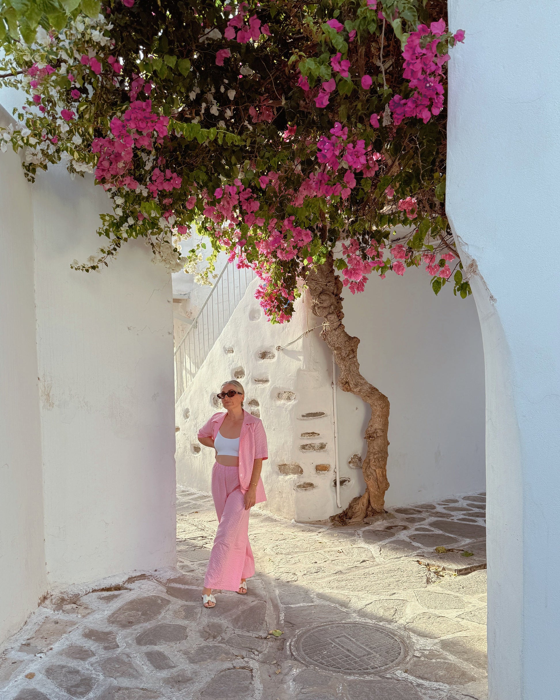 Woman in a pink dress walking through a narrow alley with white walls and pink flowers.