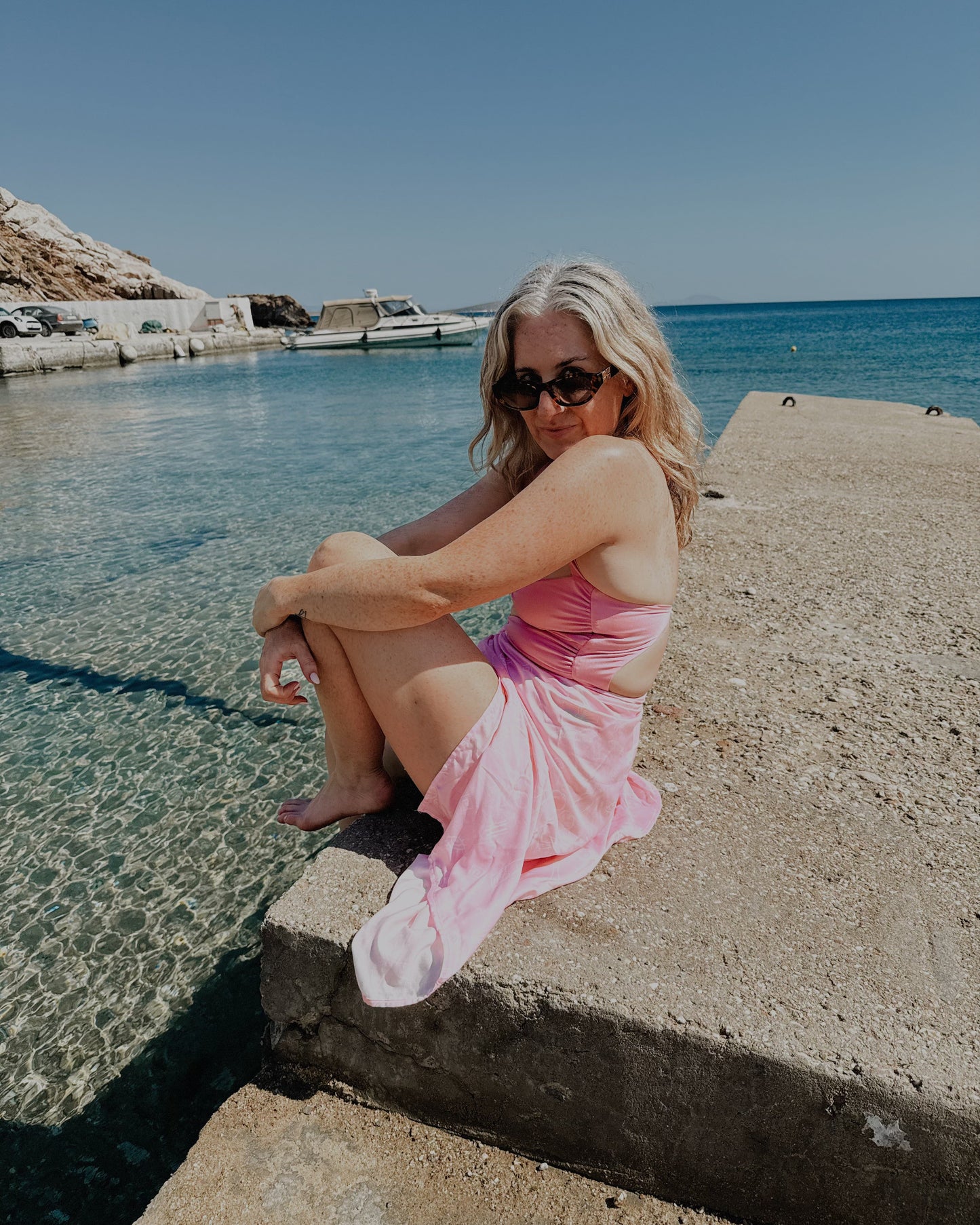 Woman in a pink dress sitting by a body of water with clear blue sky.