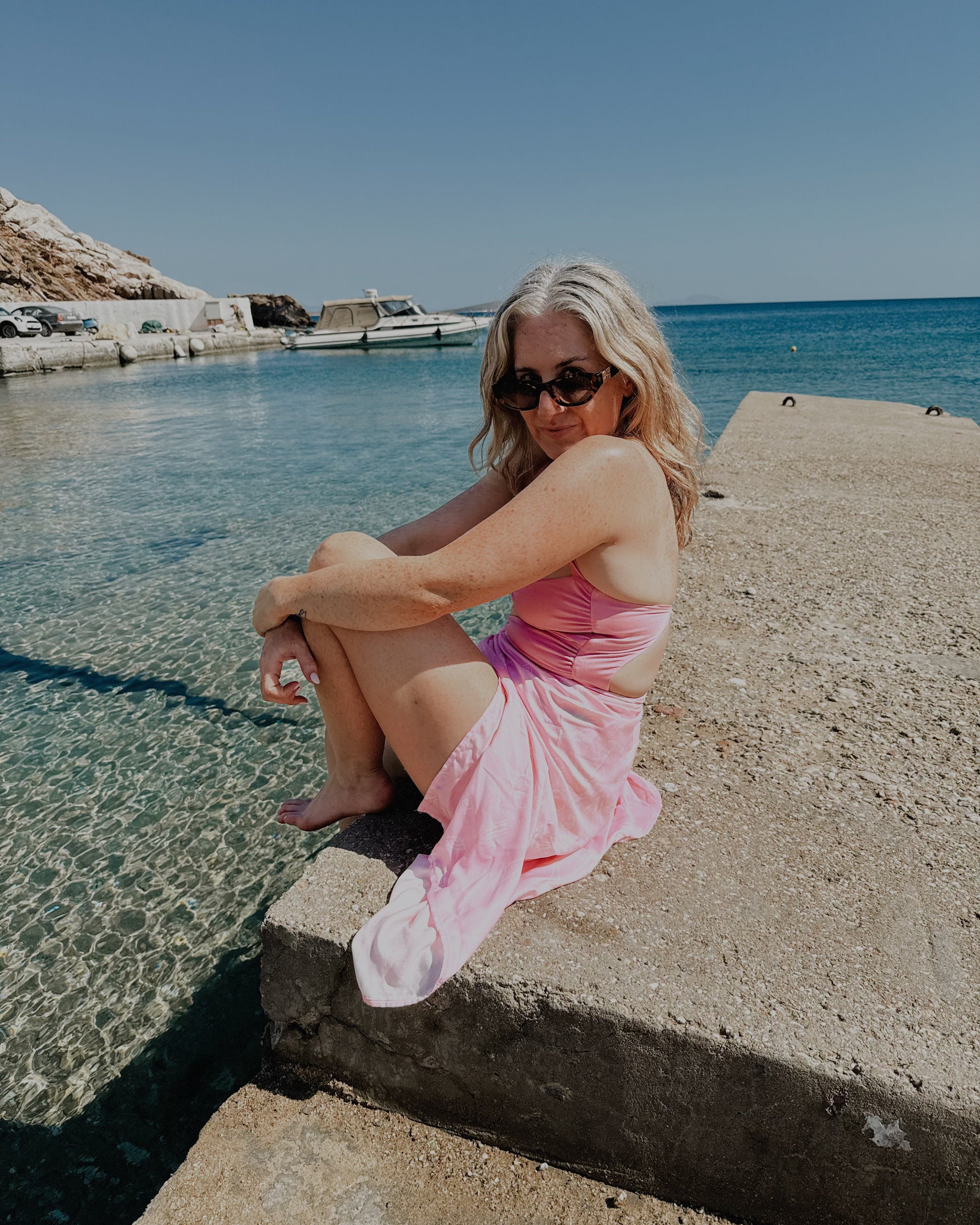 Woman in a pink dress sitting by a body of water with clear blue sky.