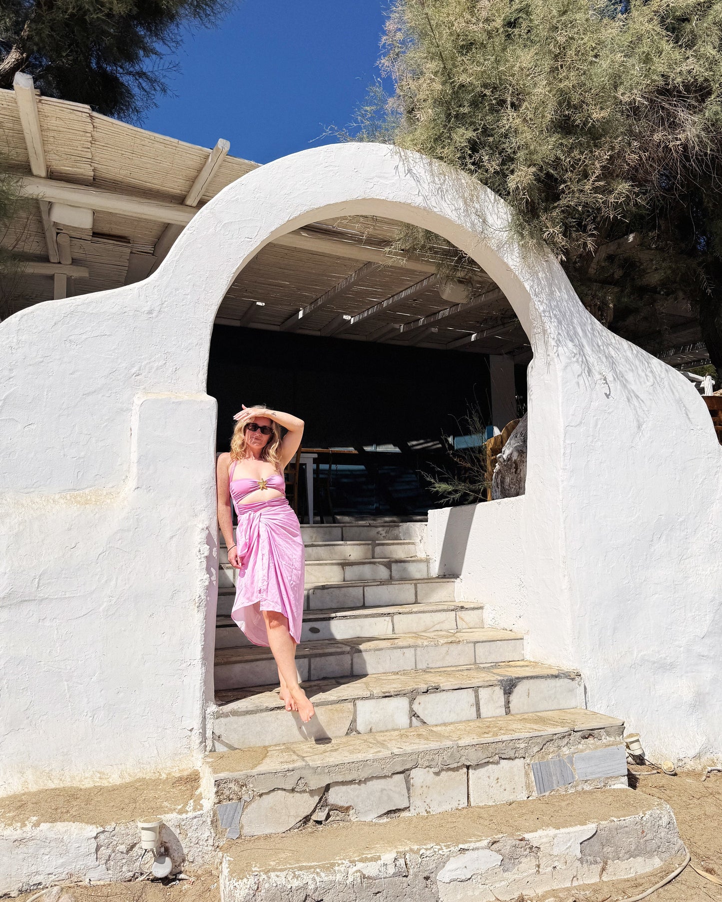 Woman in a pink dress standing on stone steps in front of a white archway with trees and blue sky in the background.