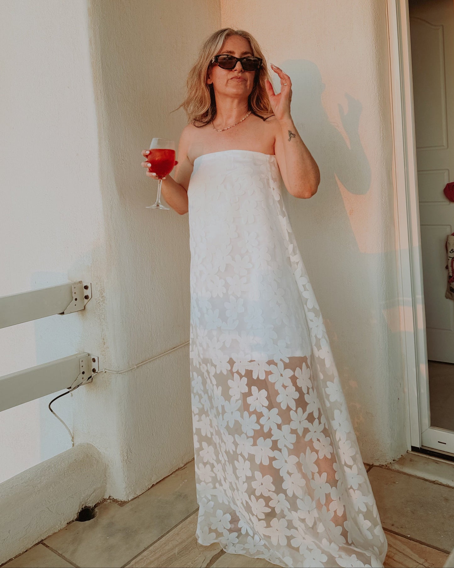 Woman in a white strapless dress holding a drink, standing in a room with a door open.