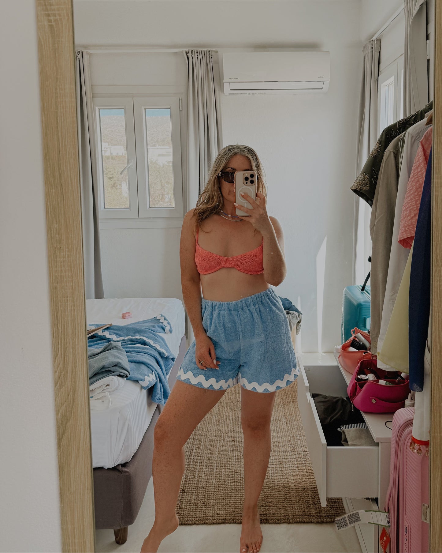 Person taking a mirror selfie wearing a red top and blue shorts in a bedroom.
