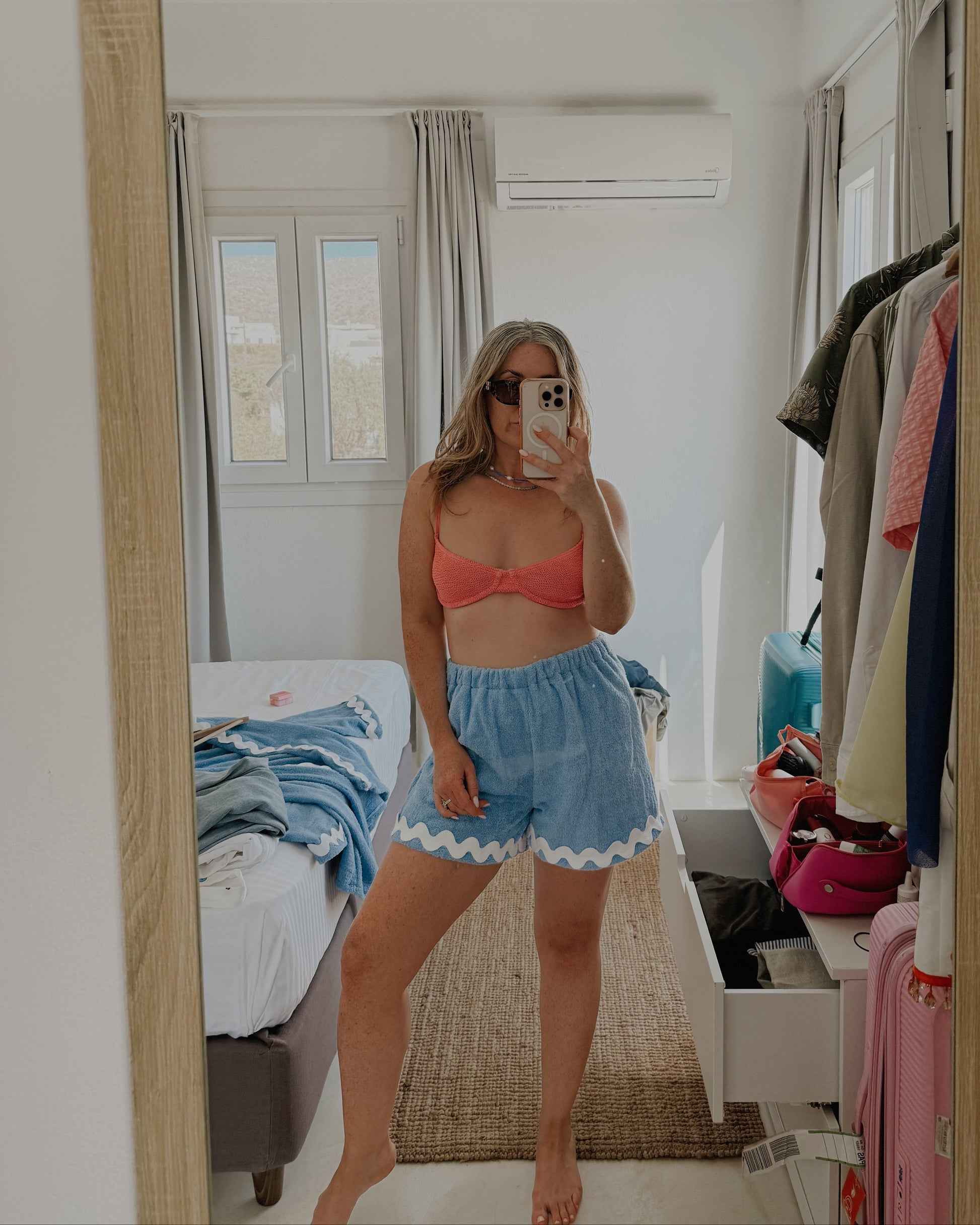 Person taking a mirror selfie wearing a red top and blue shorts in a bedroom.