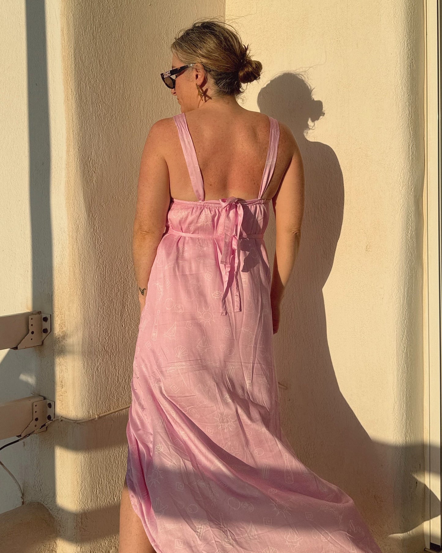 Woman in a pink dress standing by a wall with a shadow, sunlight casting a warm glow.