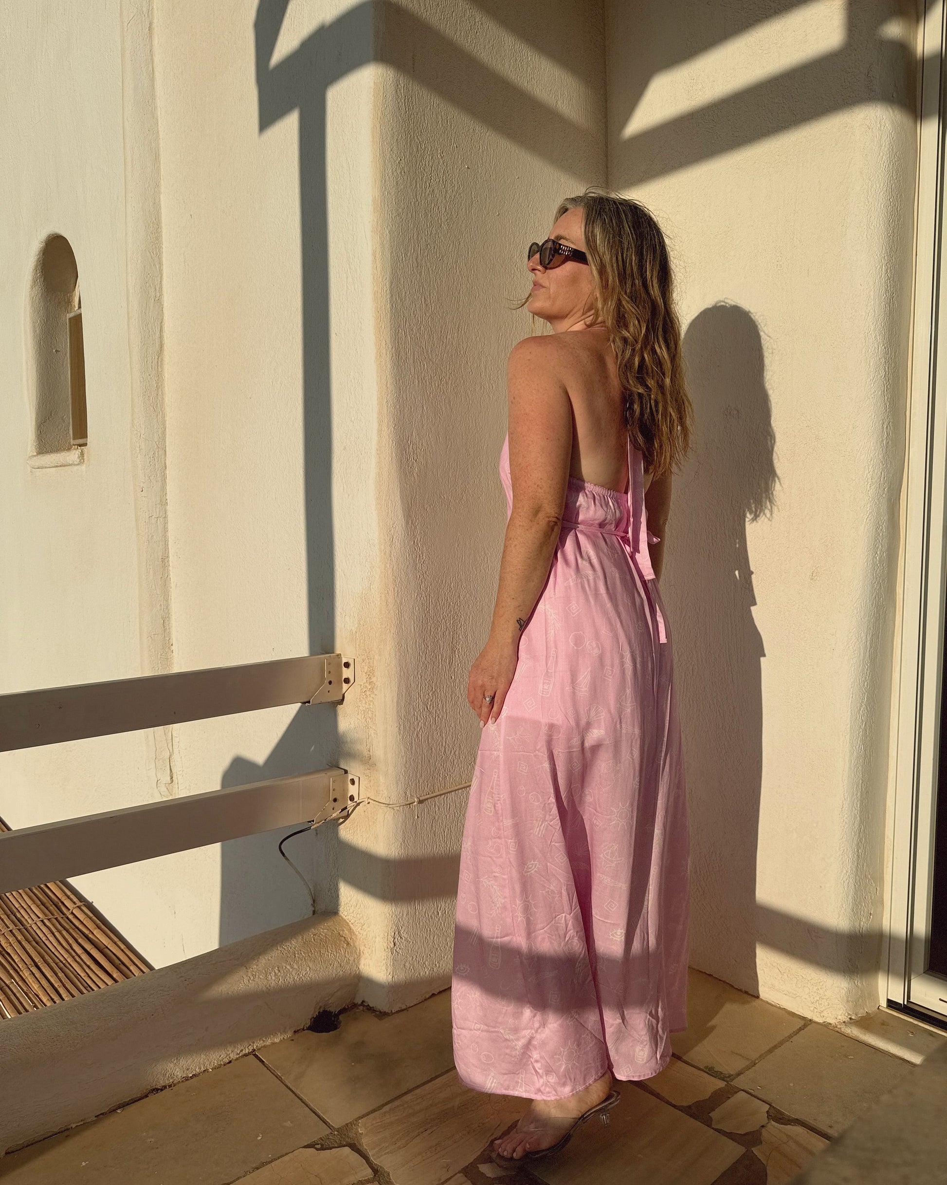 Woman in a pink dress standing by a white wall with sunlight casting shadows.