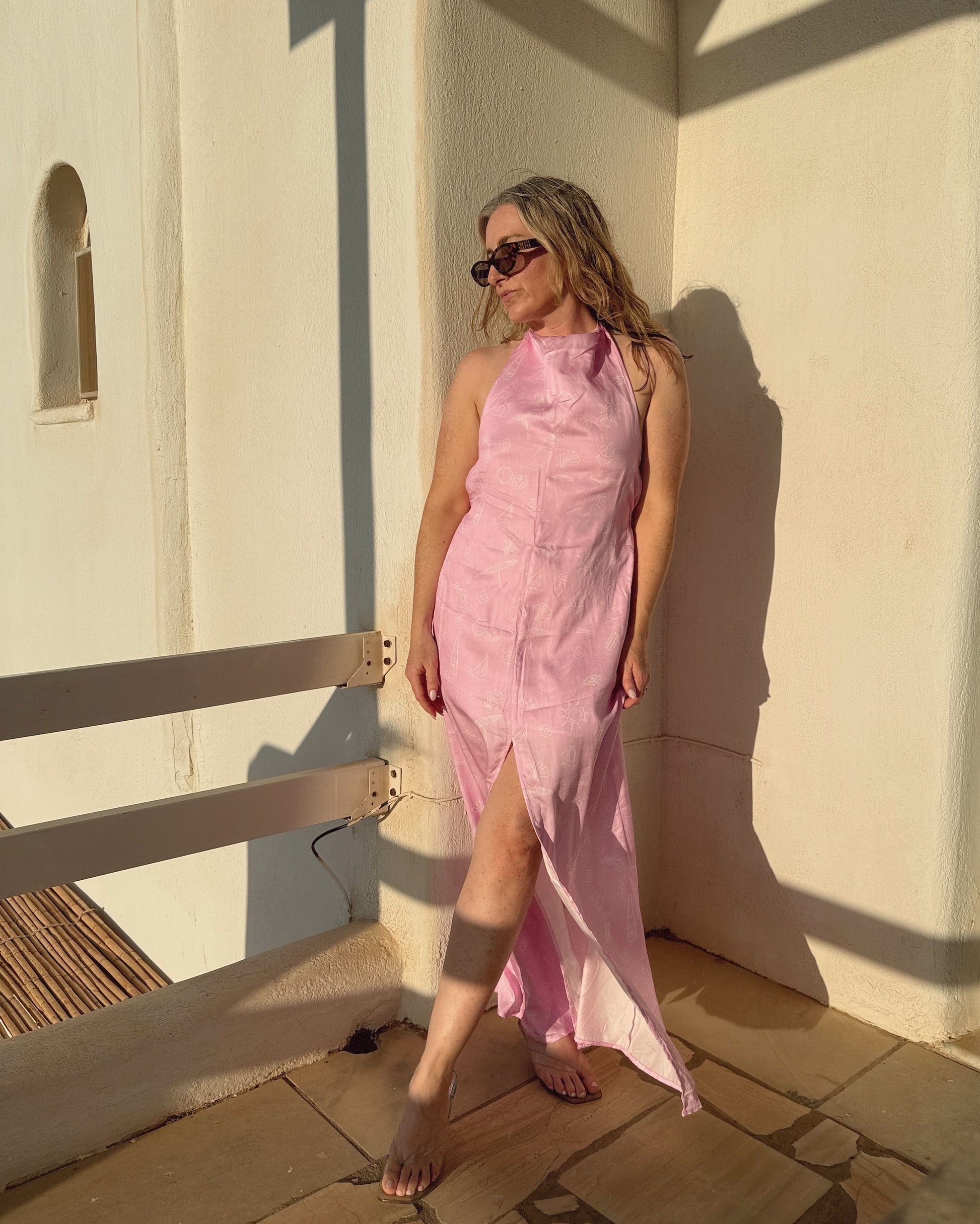 Woman in a pink dress standing against a white wall with sunlight casting shadows.