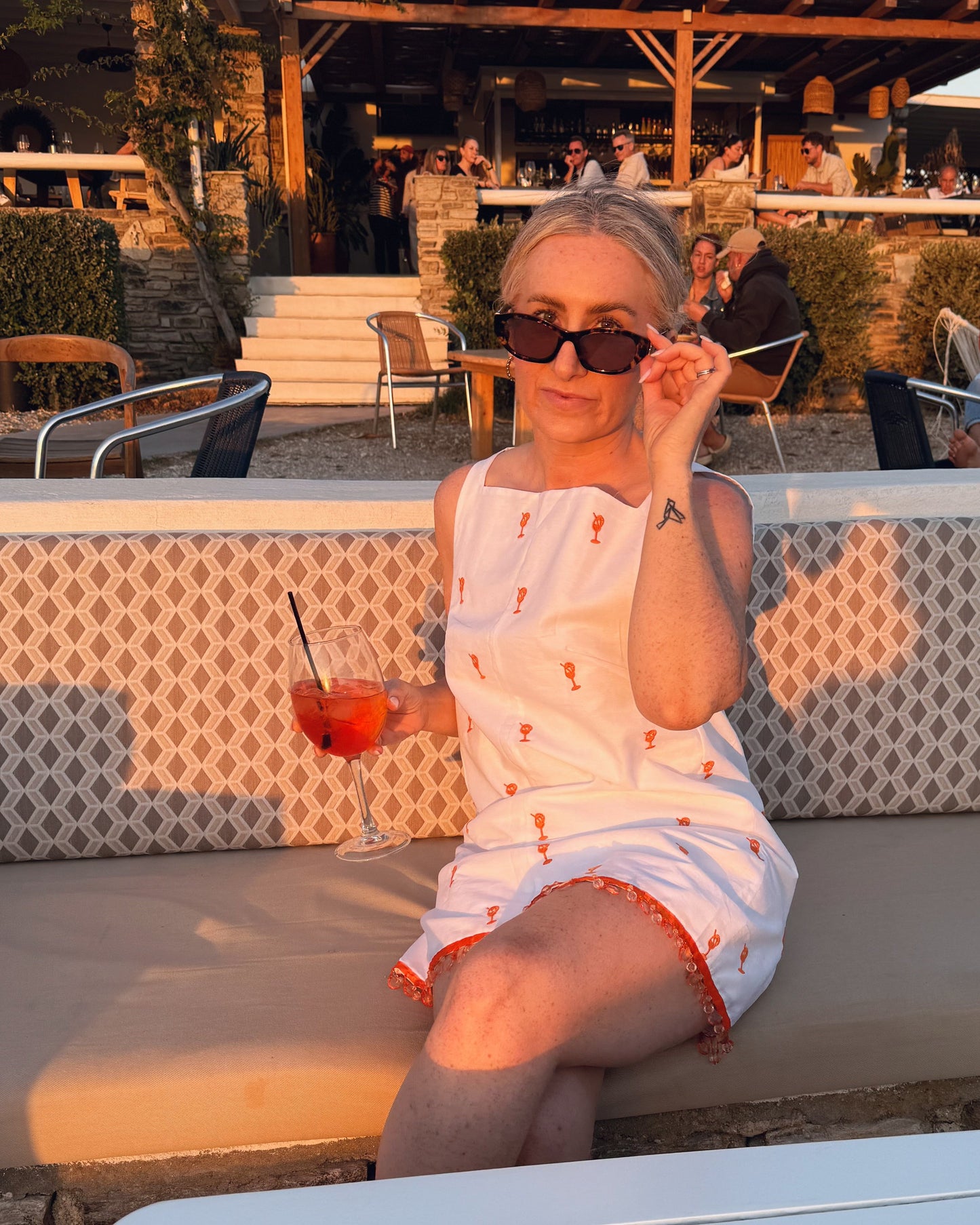 Woman sitting on a patio with a drink, surrounded by outdoor furniture and people.