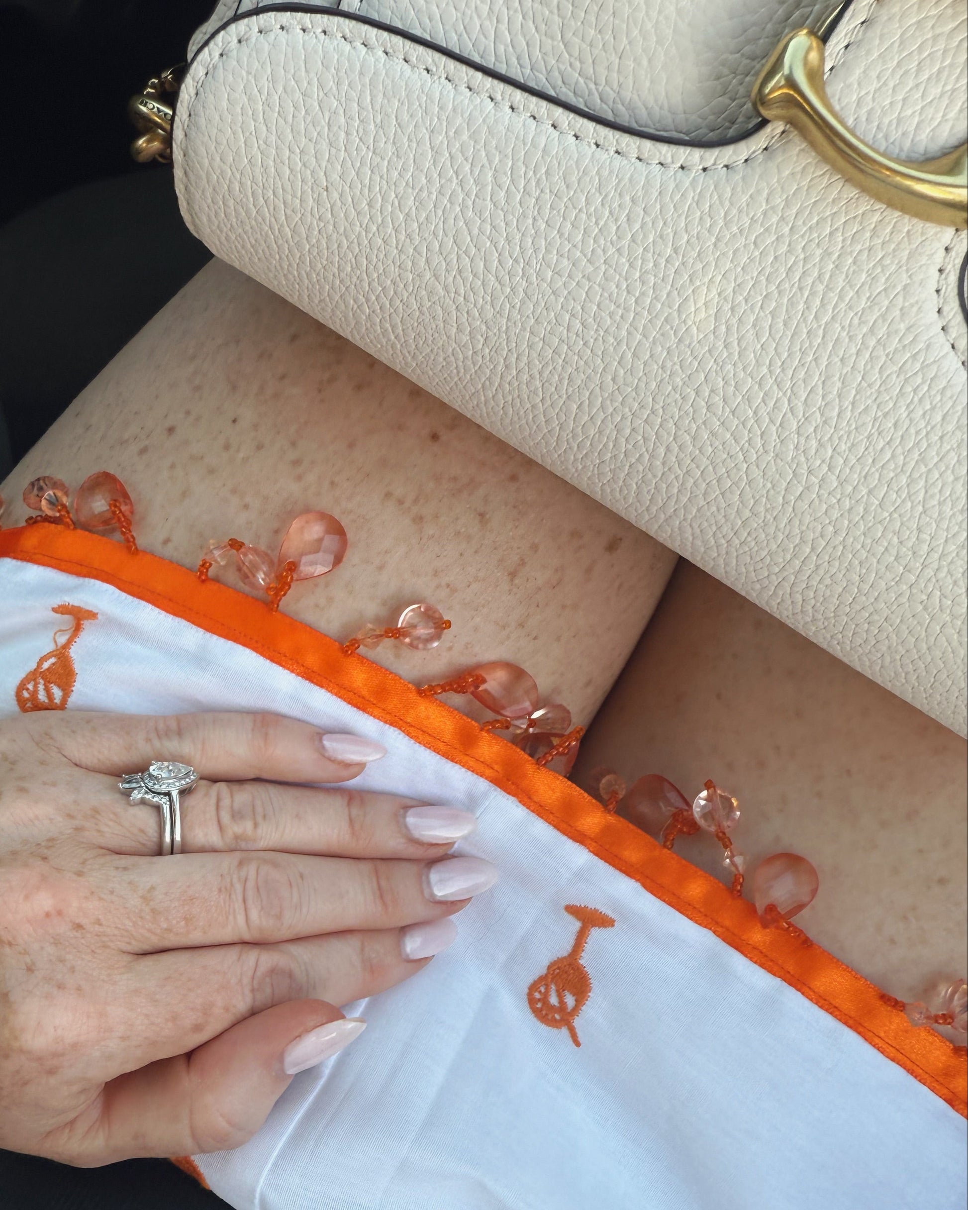Person wearing a ring on an orange and white garment with a beige handbag in the background.